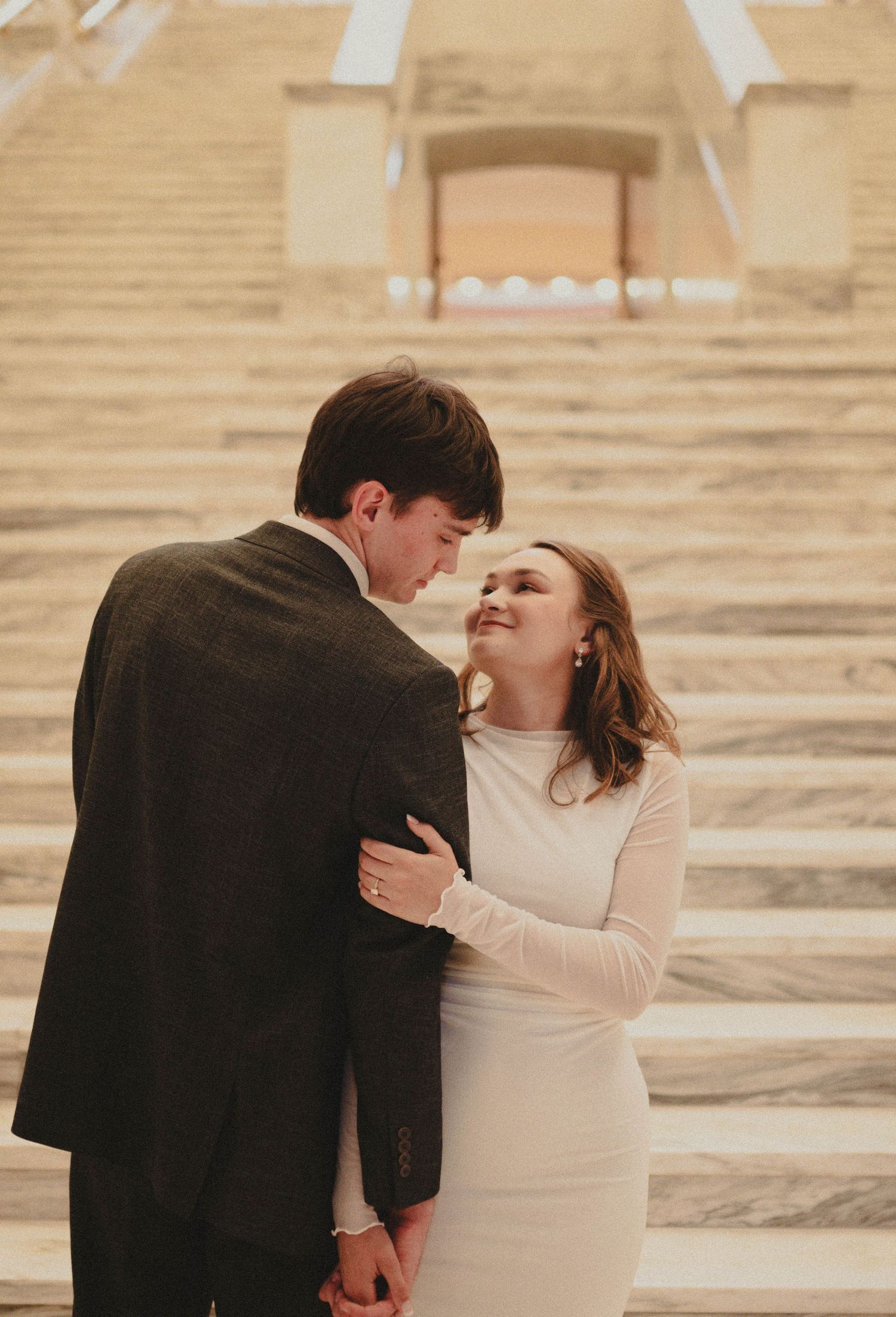 A couple shares a romantic moment on marble steps, the woman in a white dress smiling up at her partner in a gray suit.