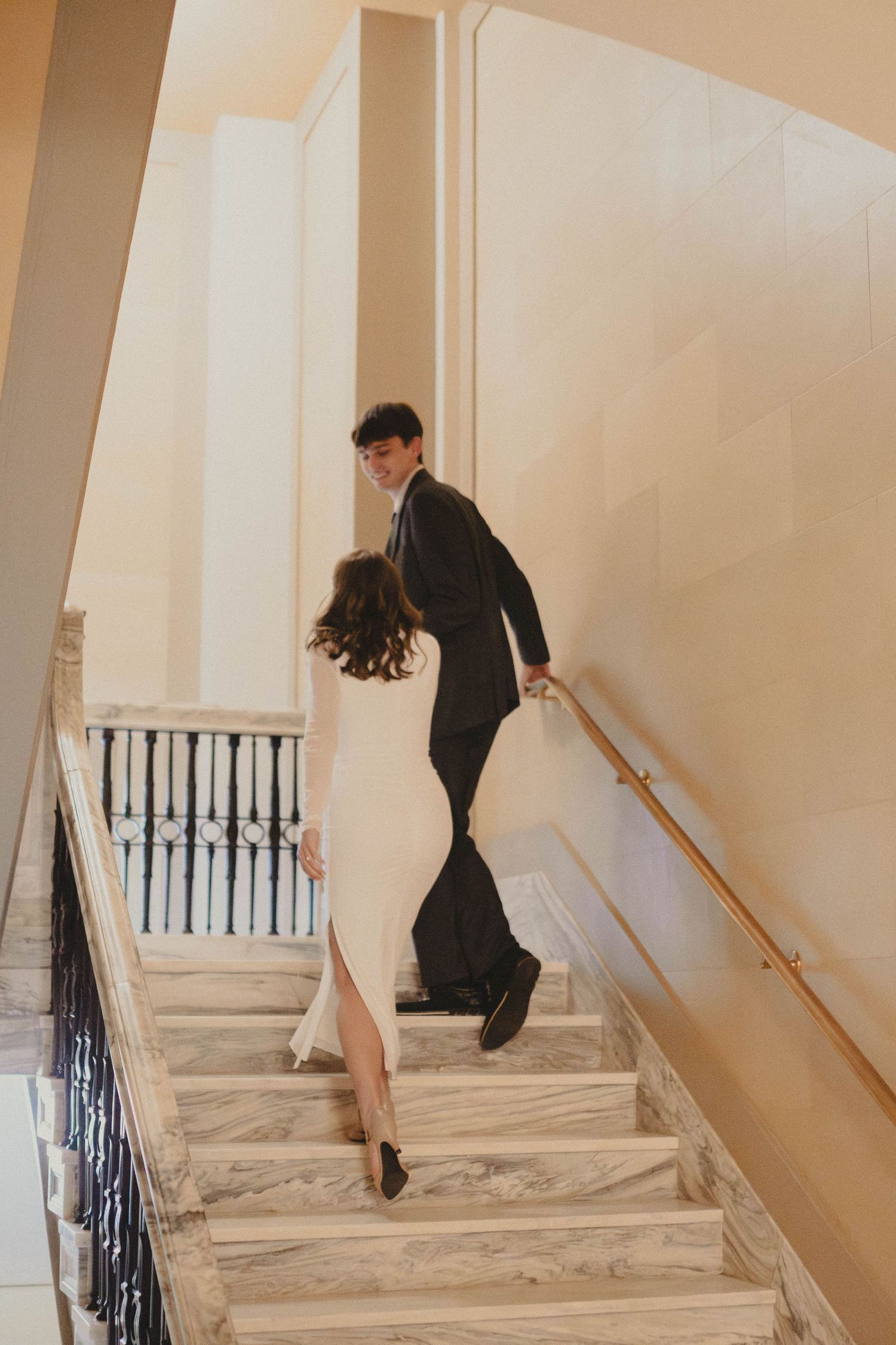 Couple walking up elegant marble staircase, woman in white dress and man in black suit, in a grand interior building.