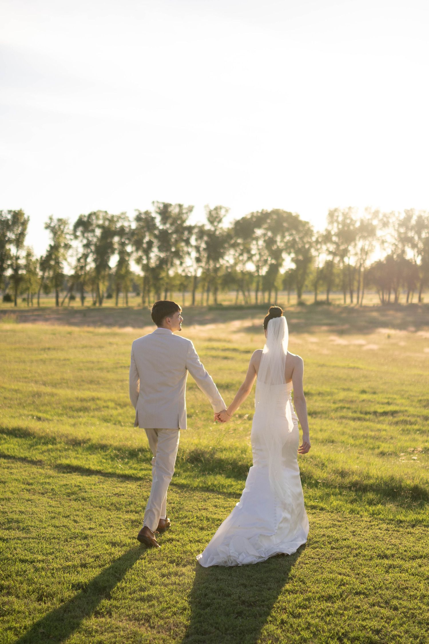 A newlywed couple walks hand in hand through a sunlit field at golden hour.