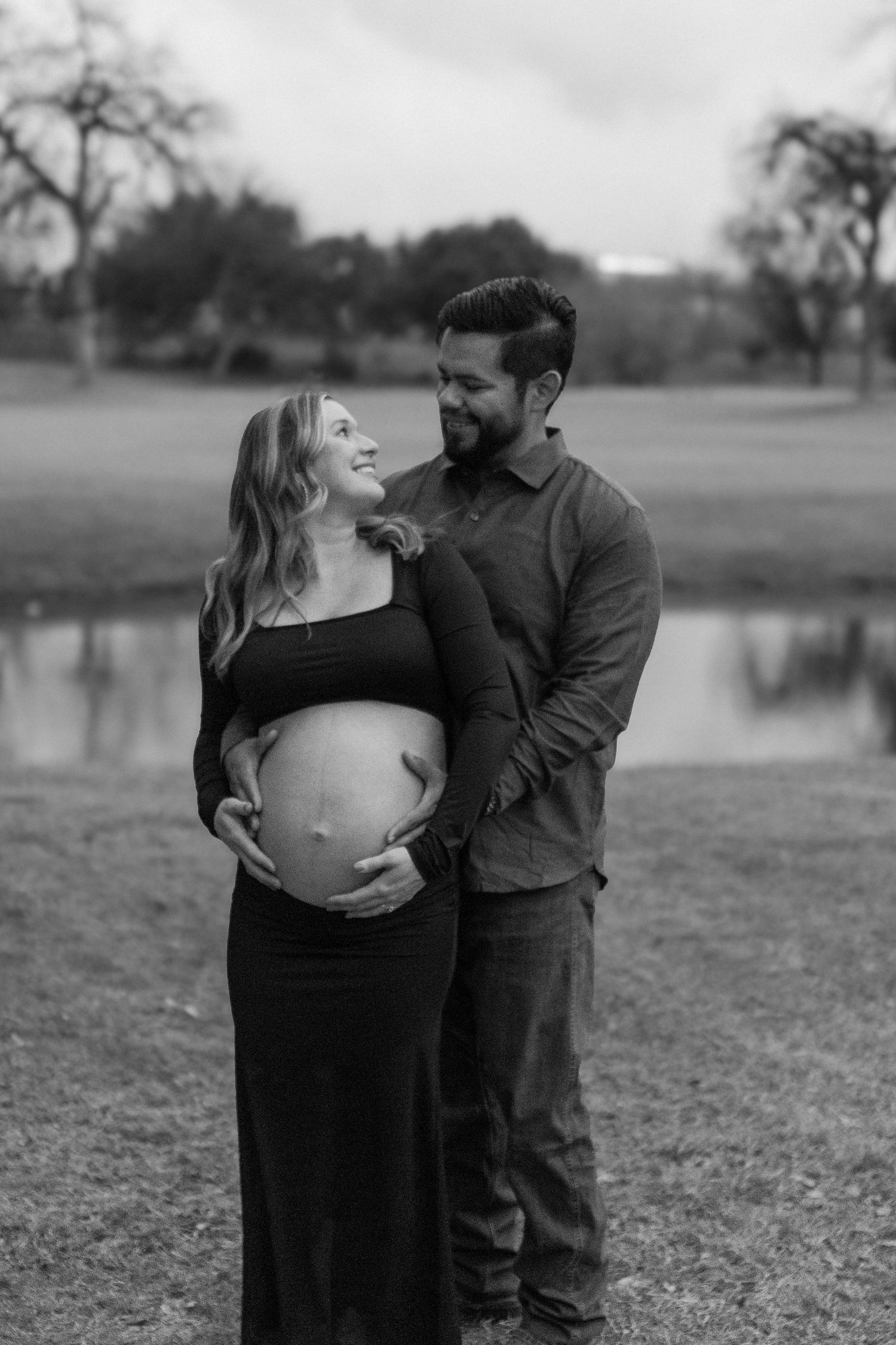Black and white maternity photo of a couple embracing near a serene lake.