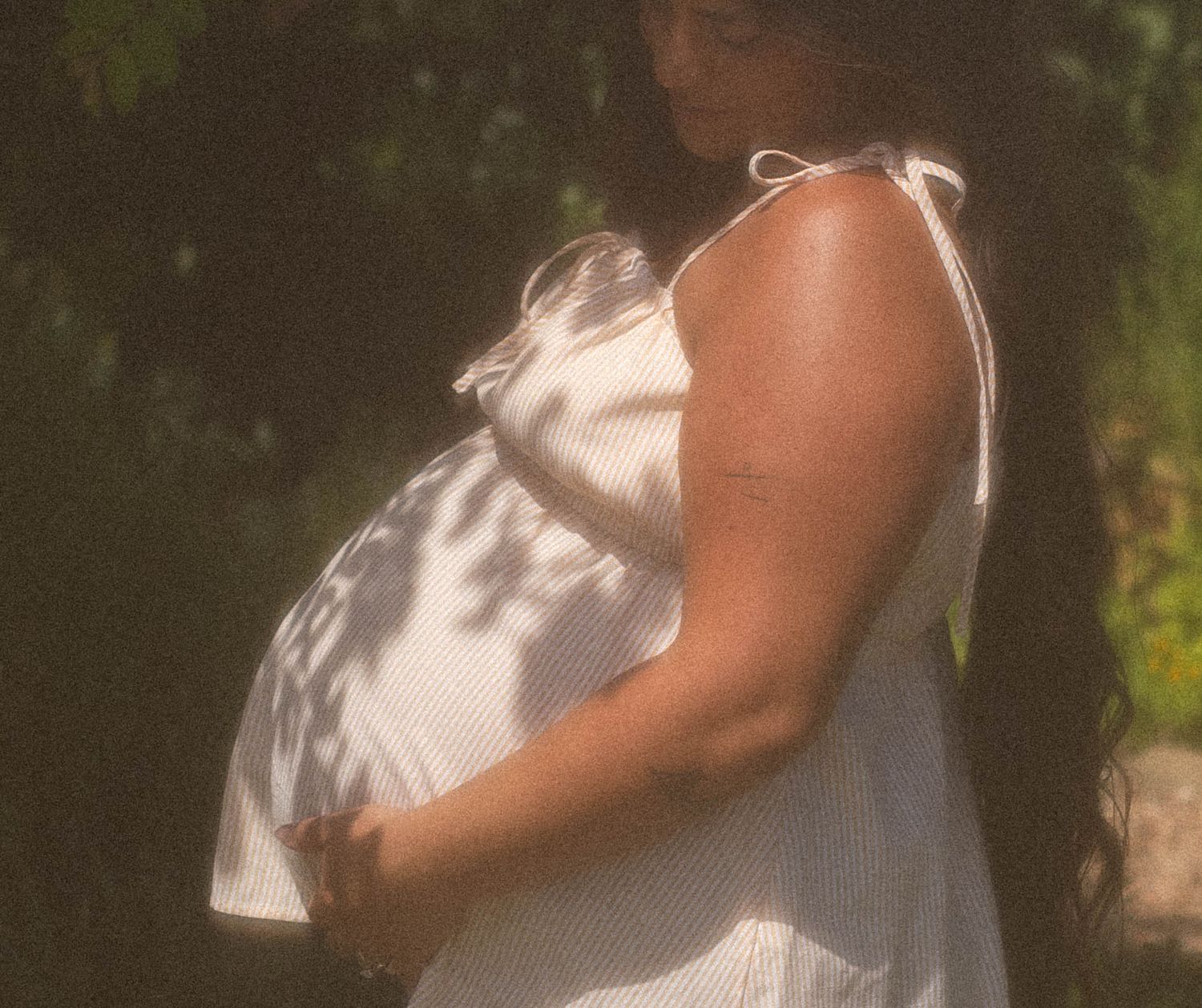 Pregnant woman in white sundress cradling baby bump outdoors in natural sunlight.