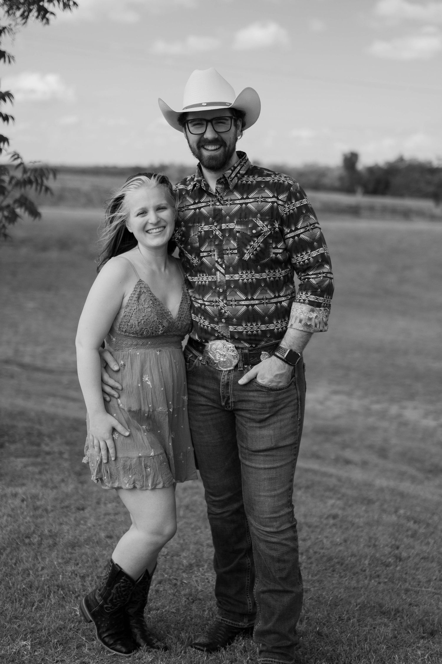 A couple poses together outdoors in western-style clothing on a grassy hillside.