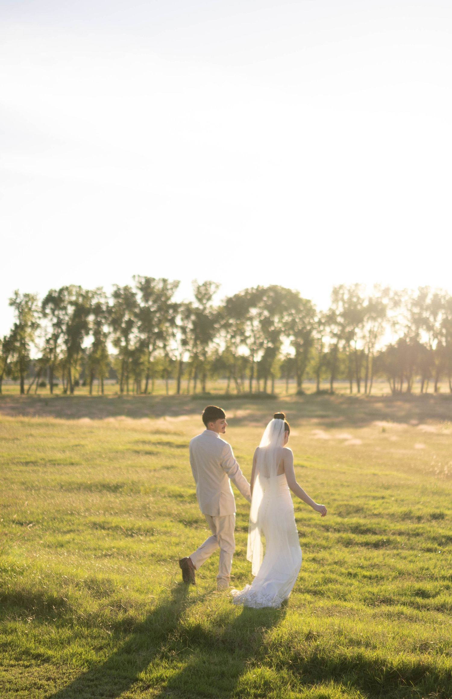 Wedding couple walks together in an open field with golden sunlight filtering through distant trees.