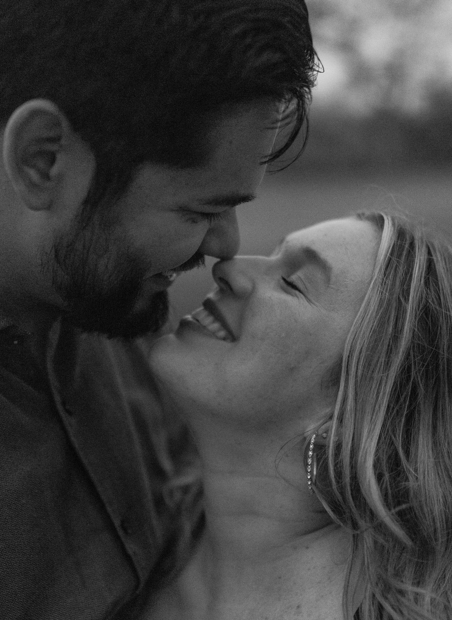 Black and white photo of a smiling couple about to kiss, captured in a romantic and intimate outdoor moment.