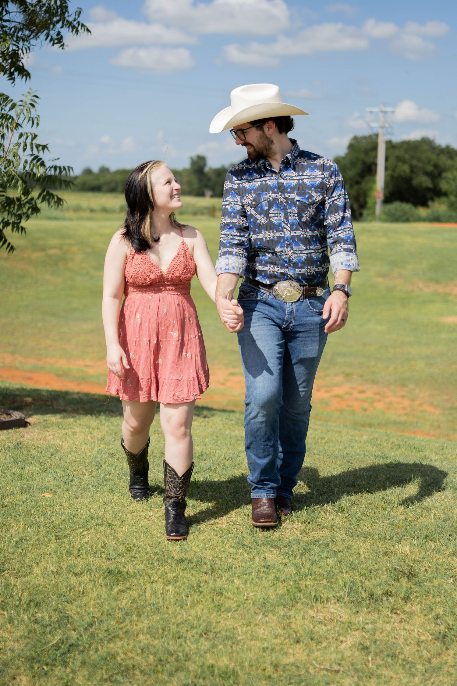A couple wearing boots stands in a sunny field with wildflowers and grass.