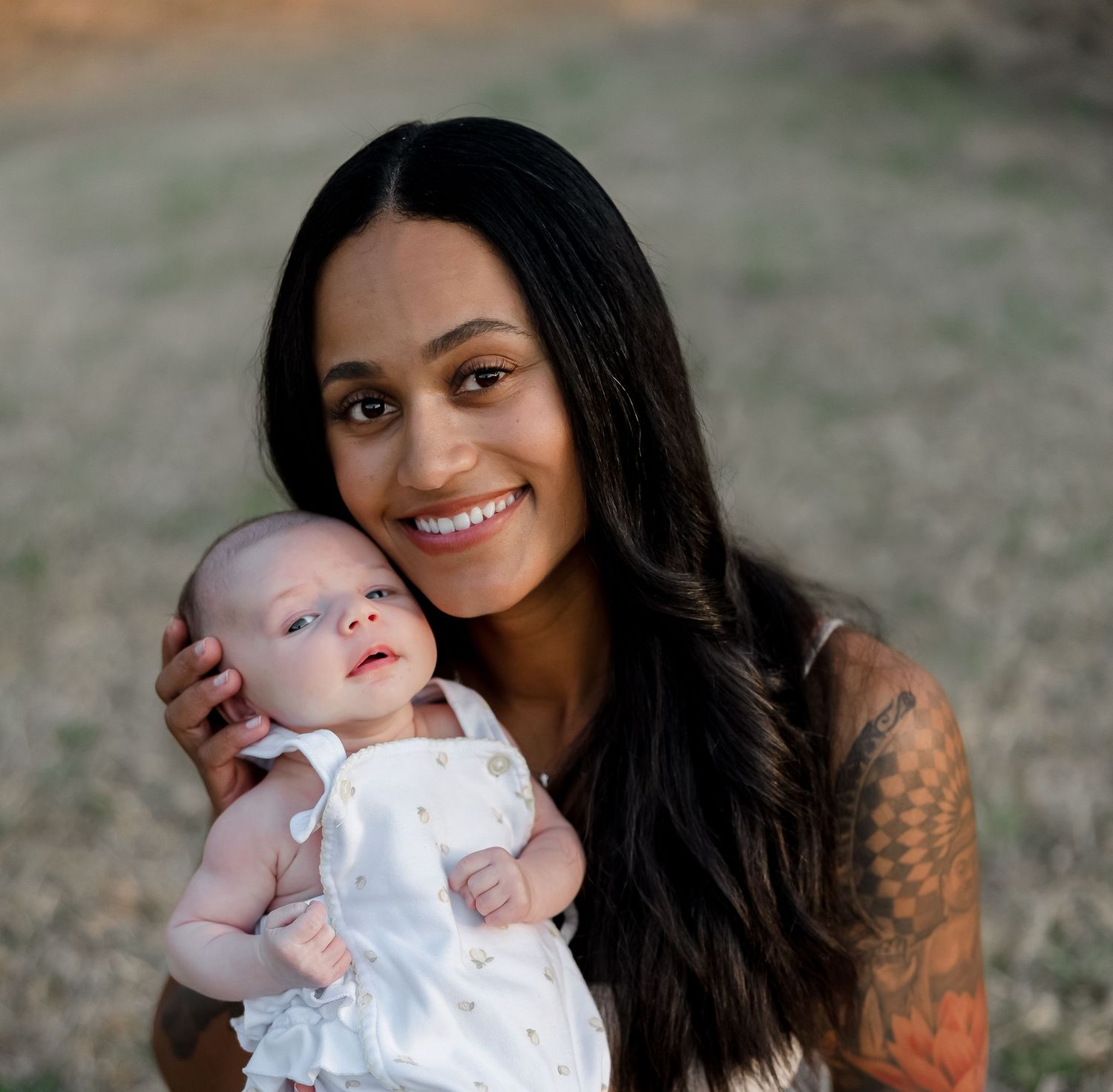 Smiling young woman with long dark hair and tattoos holding a newborn baby in white outfit outdoors.