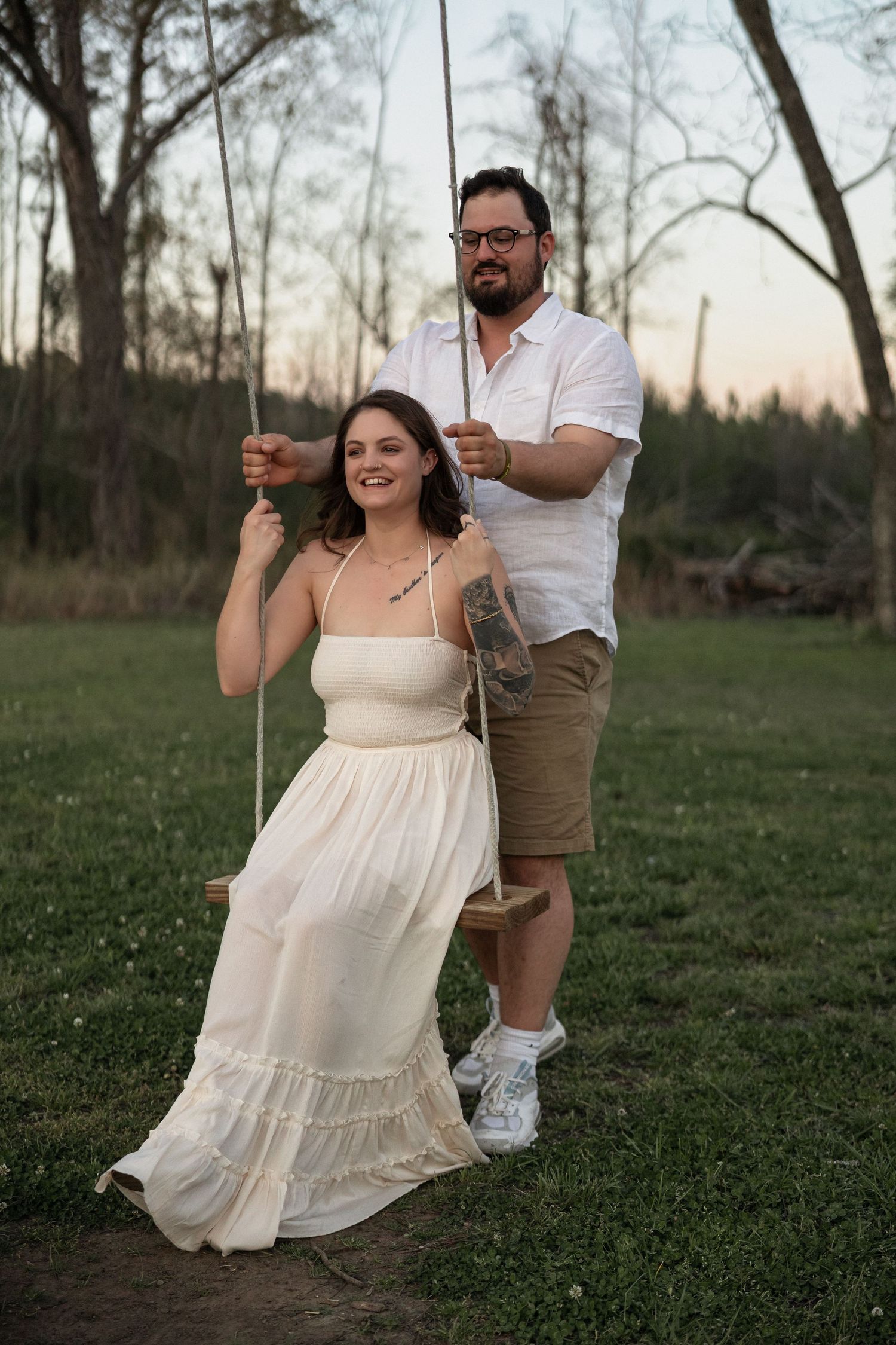 Couple posing outdoors at sunset, woman in white dress on rope swing, man in white shirt and khaki shorts pushing her.