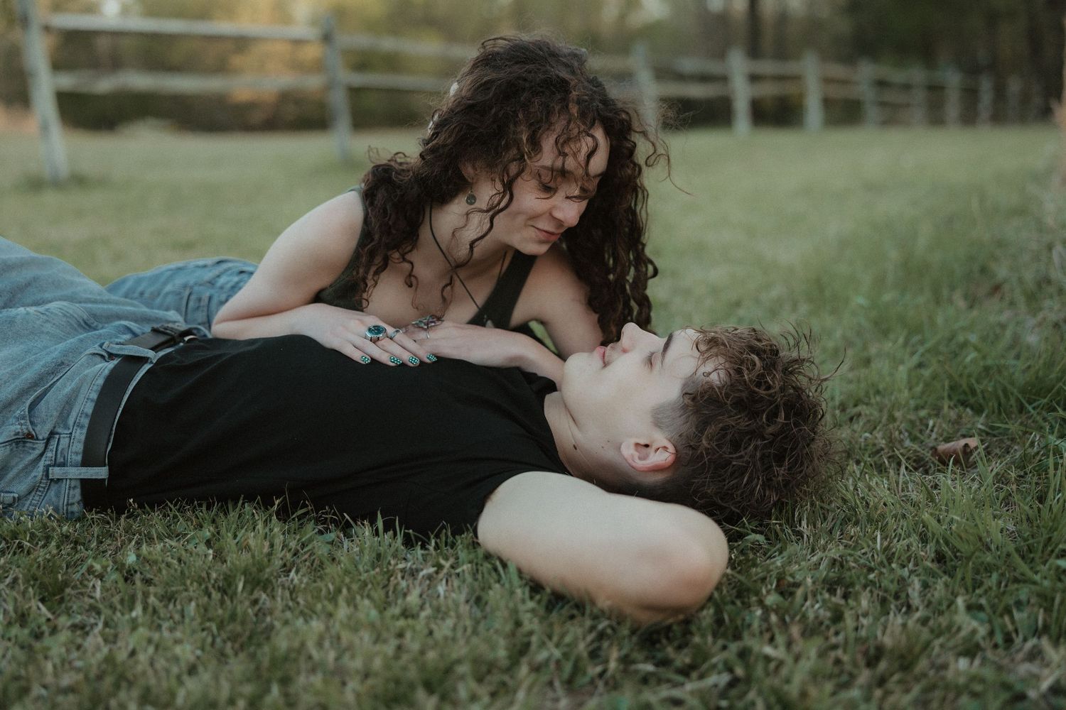 A young couple lying together on green grass in an outdoor field near a wooden fence.