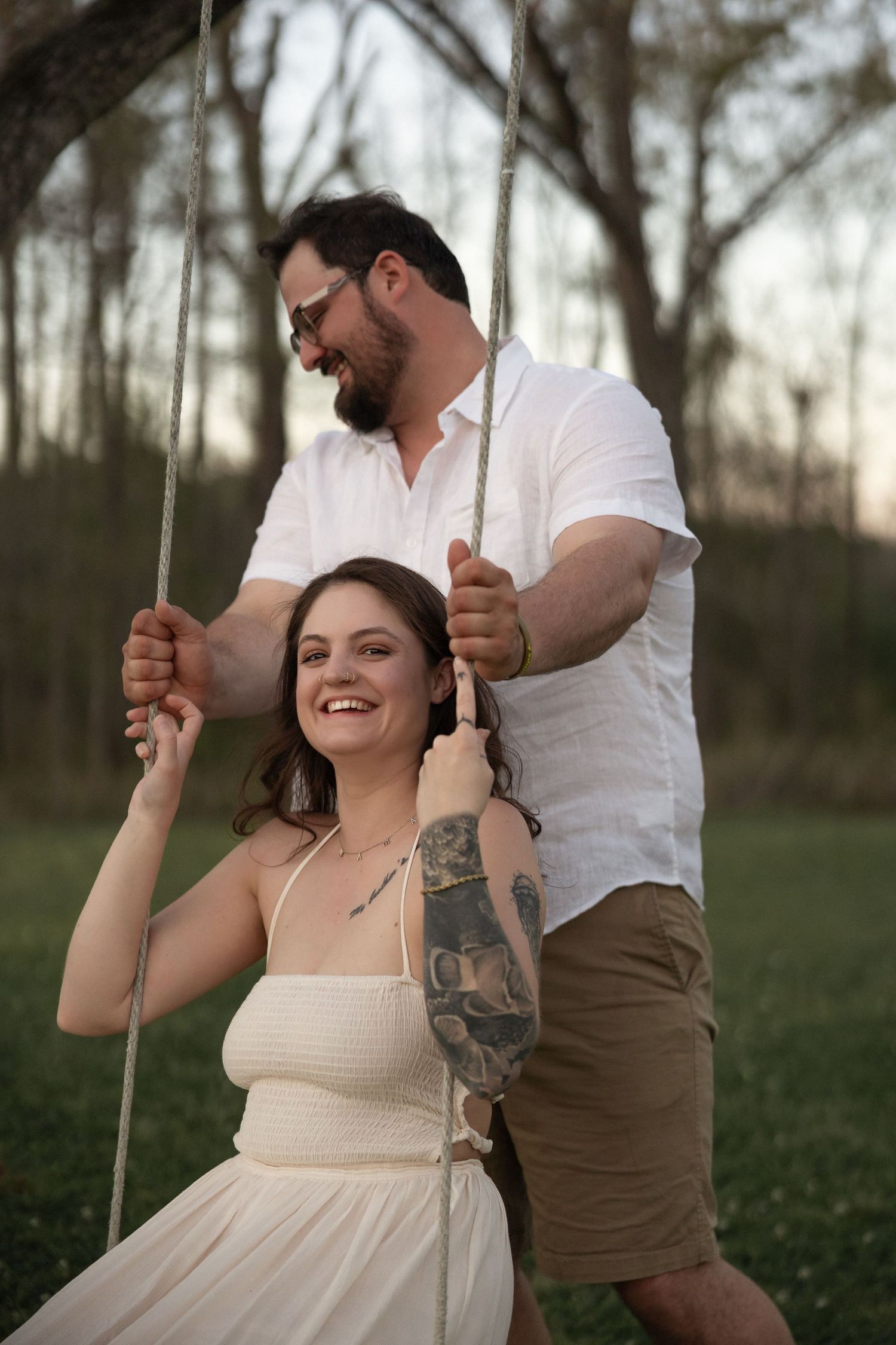 A smiling woman in a white dress sits on a rope swing while a bearded man pushes her at sunset.
