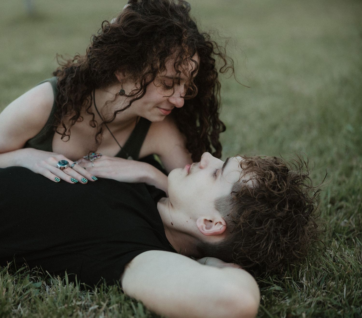 A couple shares an intimate moment on grass, woman with curly hair leaning over a young man lying down.