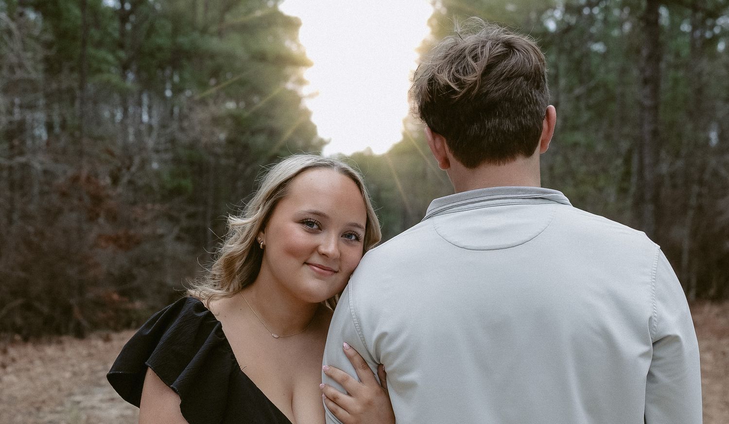 A couple in casual attire shares a romantic moment together outdoors with trees in the background.