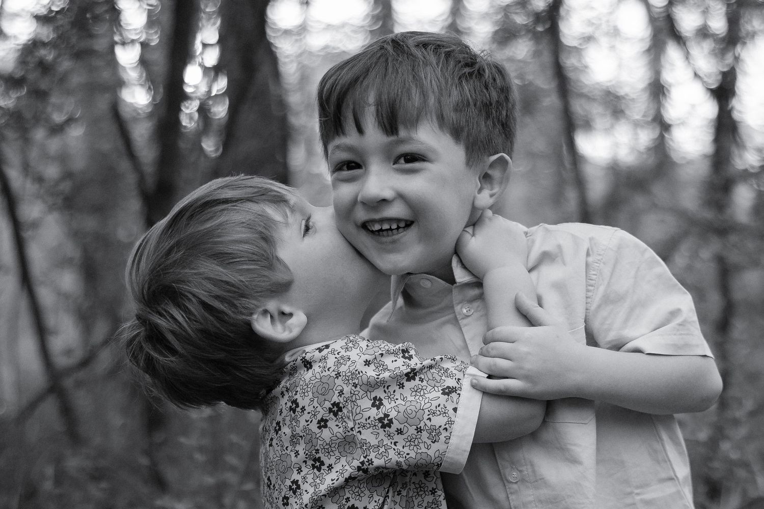 Two young children sharing a sweet moment outdoors, with a girl kissing a smiling boy on the cheek in a forest setting.