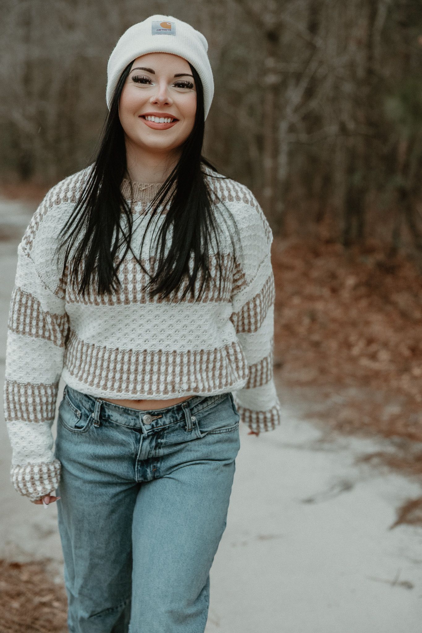 Casual winter outfit with white beanie, striped crop sweater and blue jeans on snowy forest path.