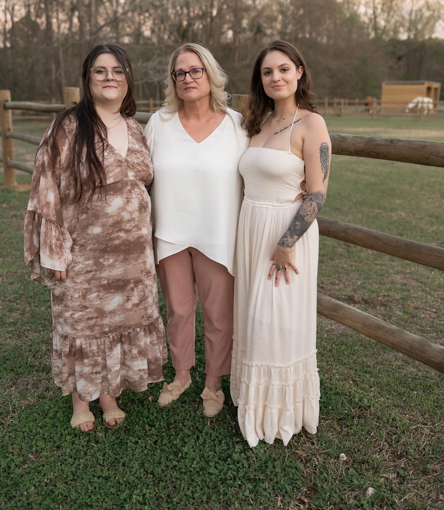 Three women posing together outdoors near a wooden fence on a green lawn wearing elegant neutral-toned dresses.