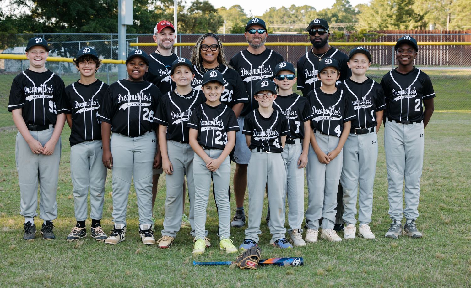 Youth baseball team poses for group photo on field, players in black uniforms with coaches standing behind them.