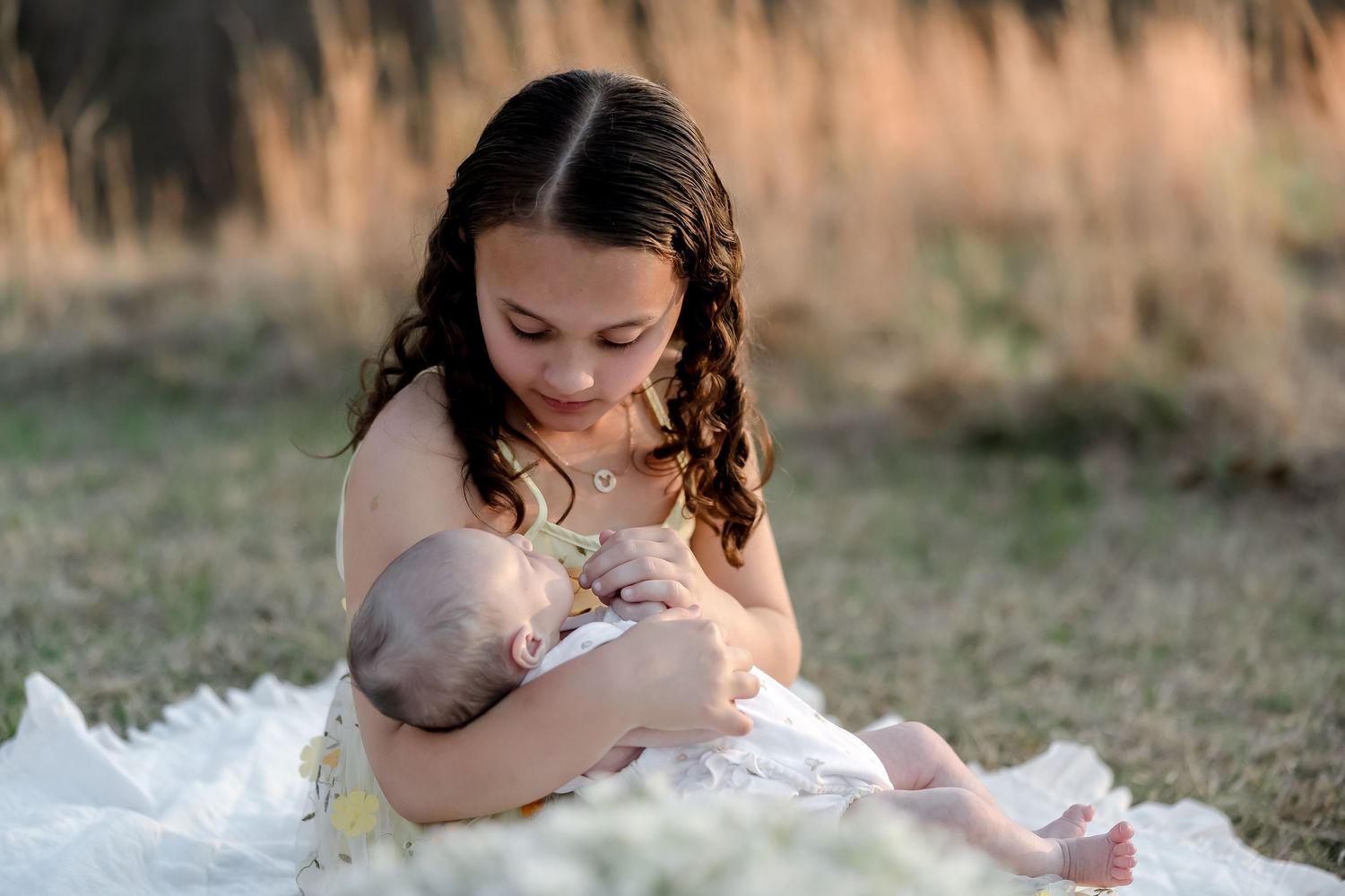 A young child in a white dress sits outdoors while cradling a baby in their arms.