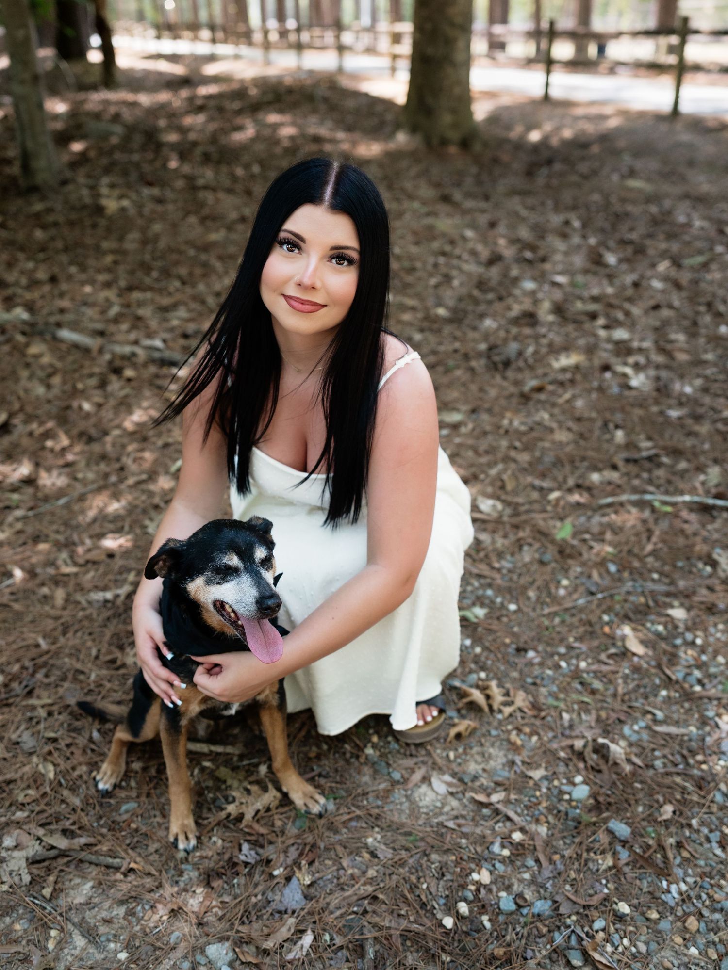Young woman in white dress crouching with a black and tan dog in a wooded outdoor setting.