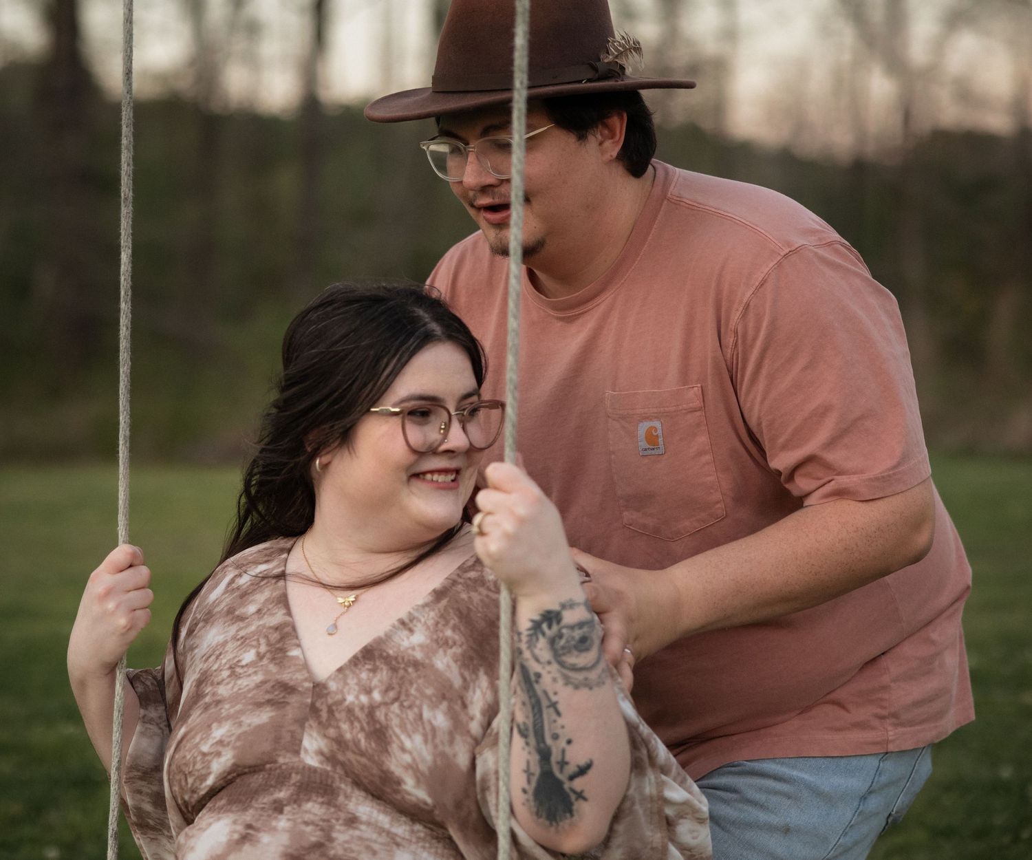 A couple smiles on a rope swing outdoors, woman in floral top and man in pink shirt and brown hat.