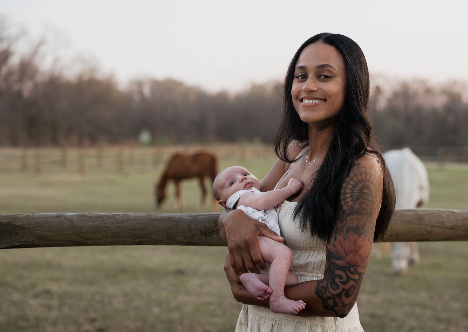 Tattooed woman smiling while holding newborn baby outdoors on a farm with horses grazing in background.