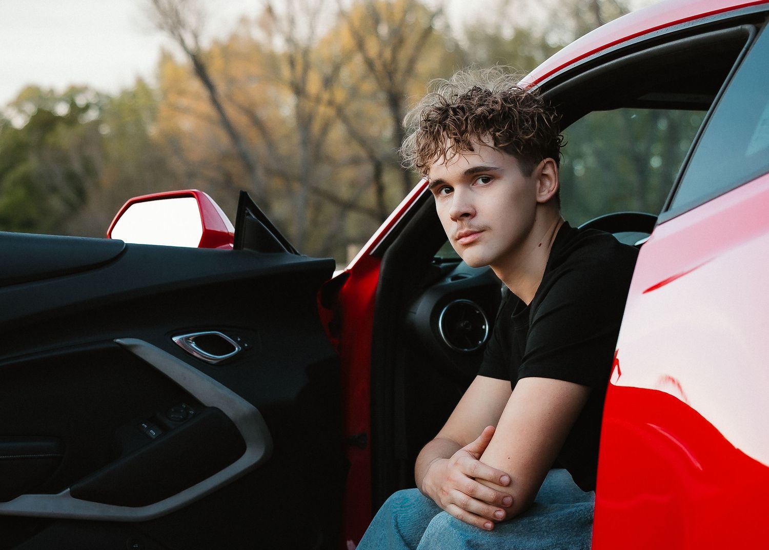 Young man with curly hair sitting in open door of red sports car outdoors, arms crossed, casual black shirt.