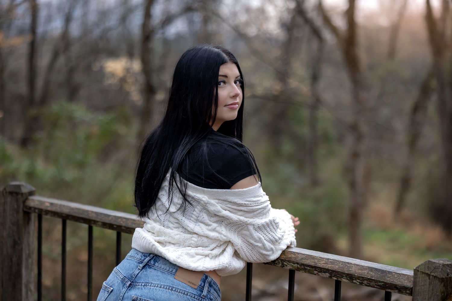 A woman in a denim jacket and white scarf looks away thoughtfully in an autumn forest setting.