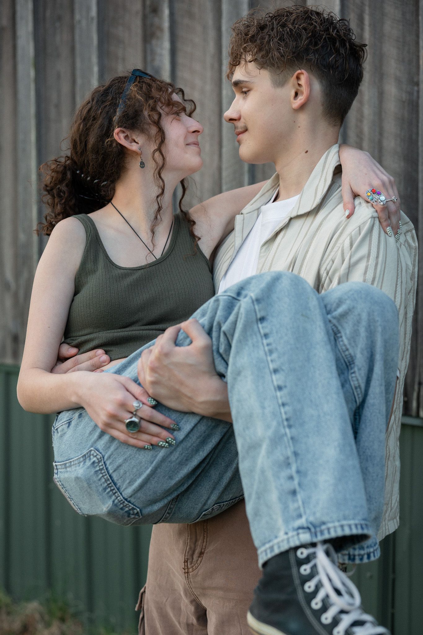 Young couple embracing and gazing at each other near a rustic wooden wall, man holding woman in his arms.