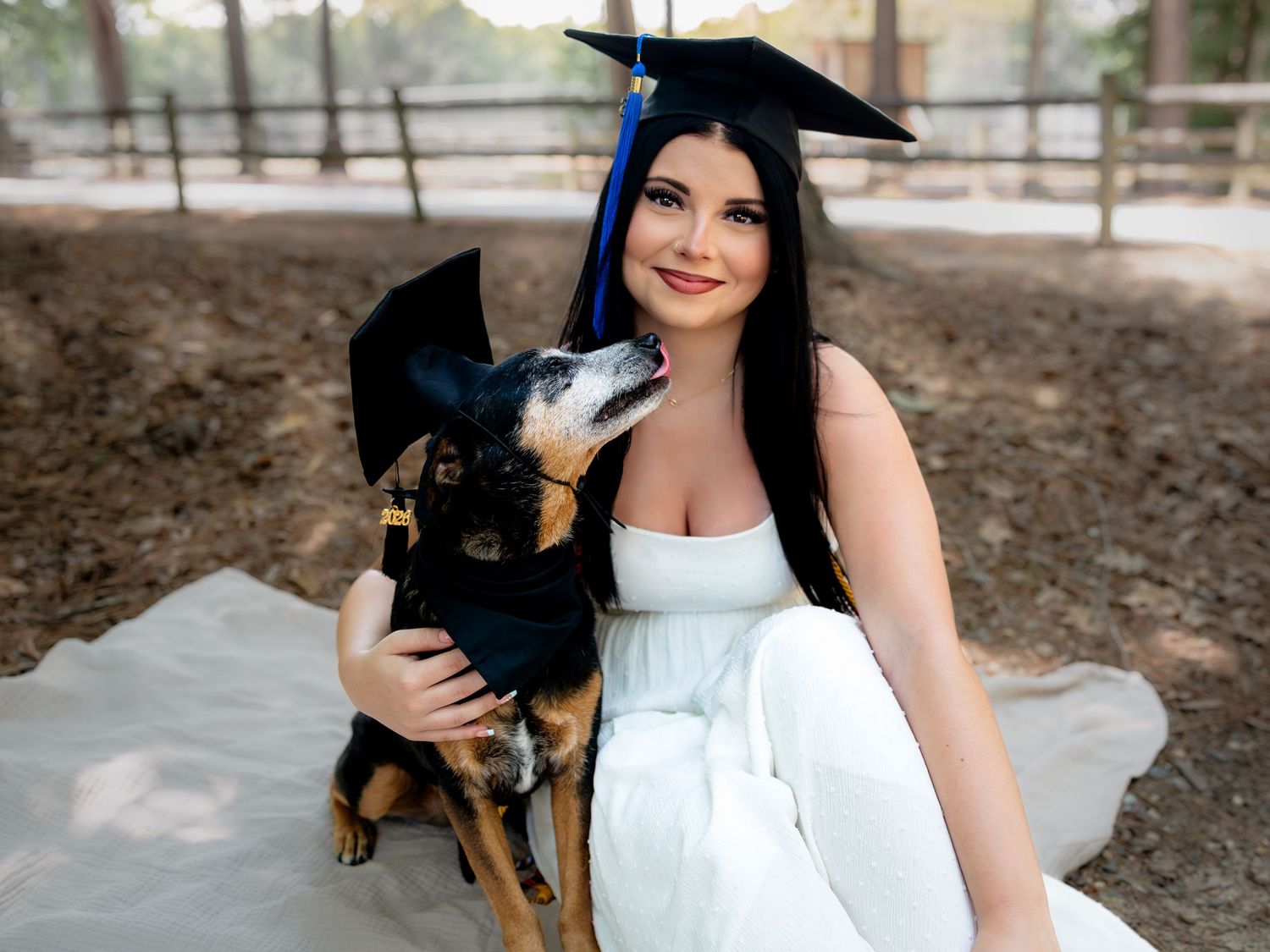 A smiling woman in a white dress and graduation cap poses with her dog also wearing a graduation cap outdoors.