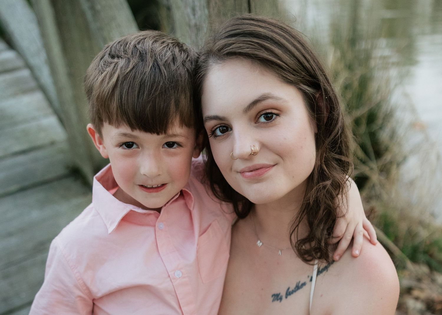 A young boy in a pink shirt smiles beside a brunette woman with tattoos during an outdoor portrait session.