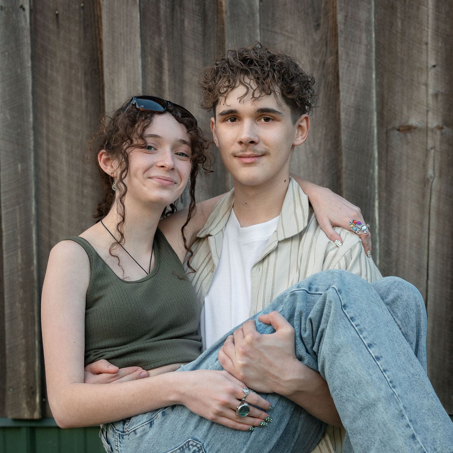 Two young people sitting together smiling against a wooden wall, with the boy's arm around the girl.