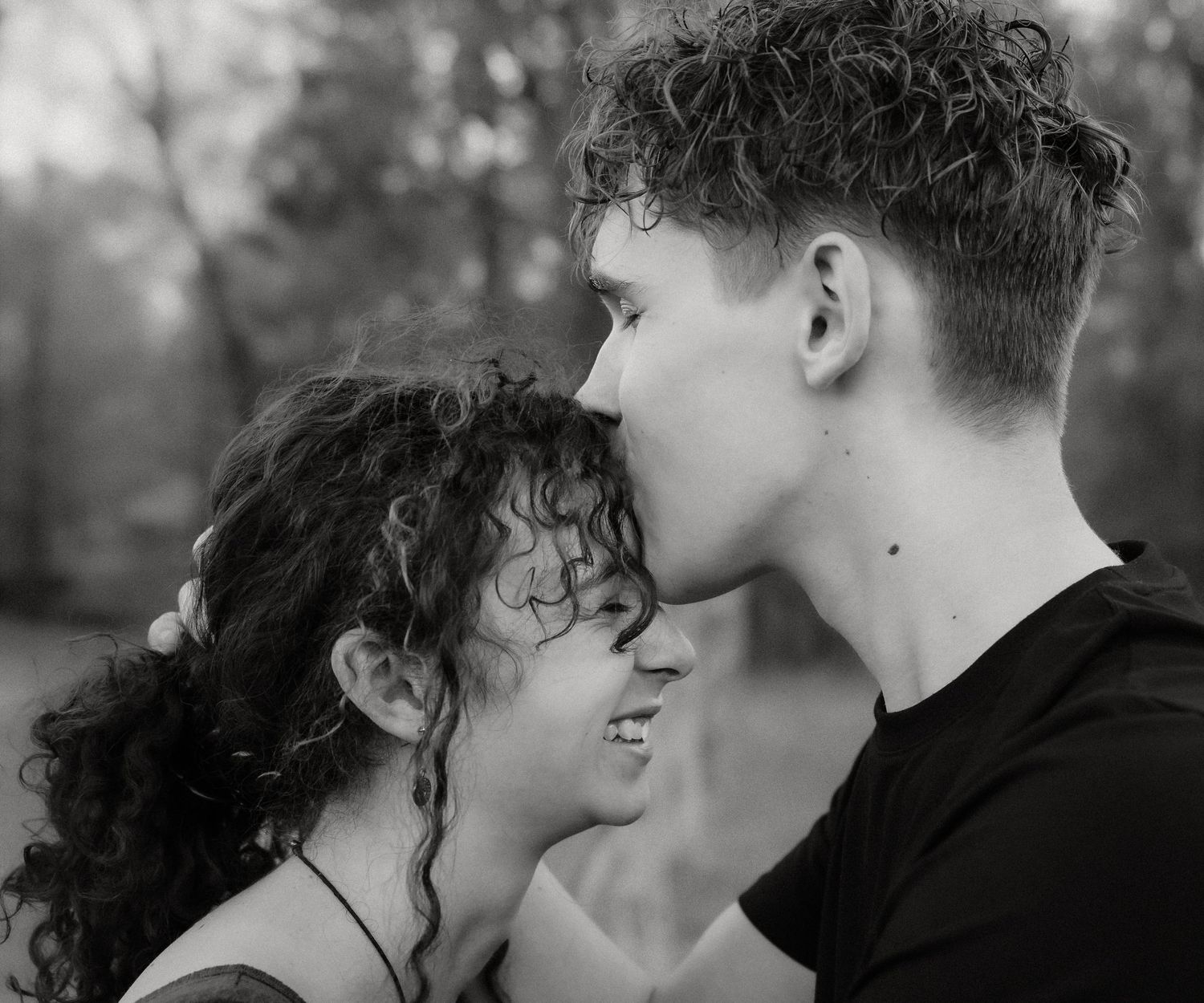 Black and white photo of a young couple sharing a tender romantic moment outdoors, boy leaning in to kiss girl.