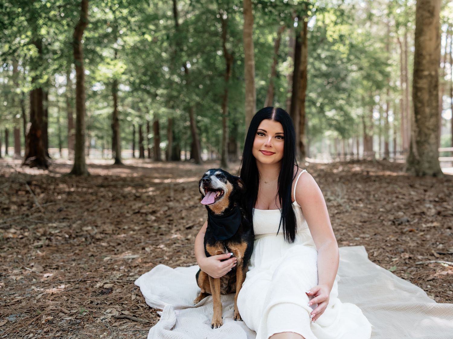 Woman in white dress sitting with her happy dog in a serene wooded forest setting.