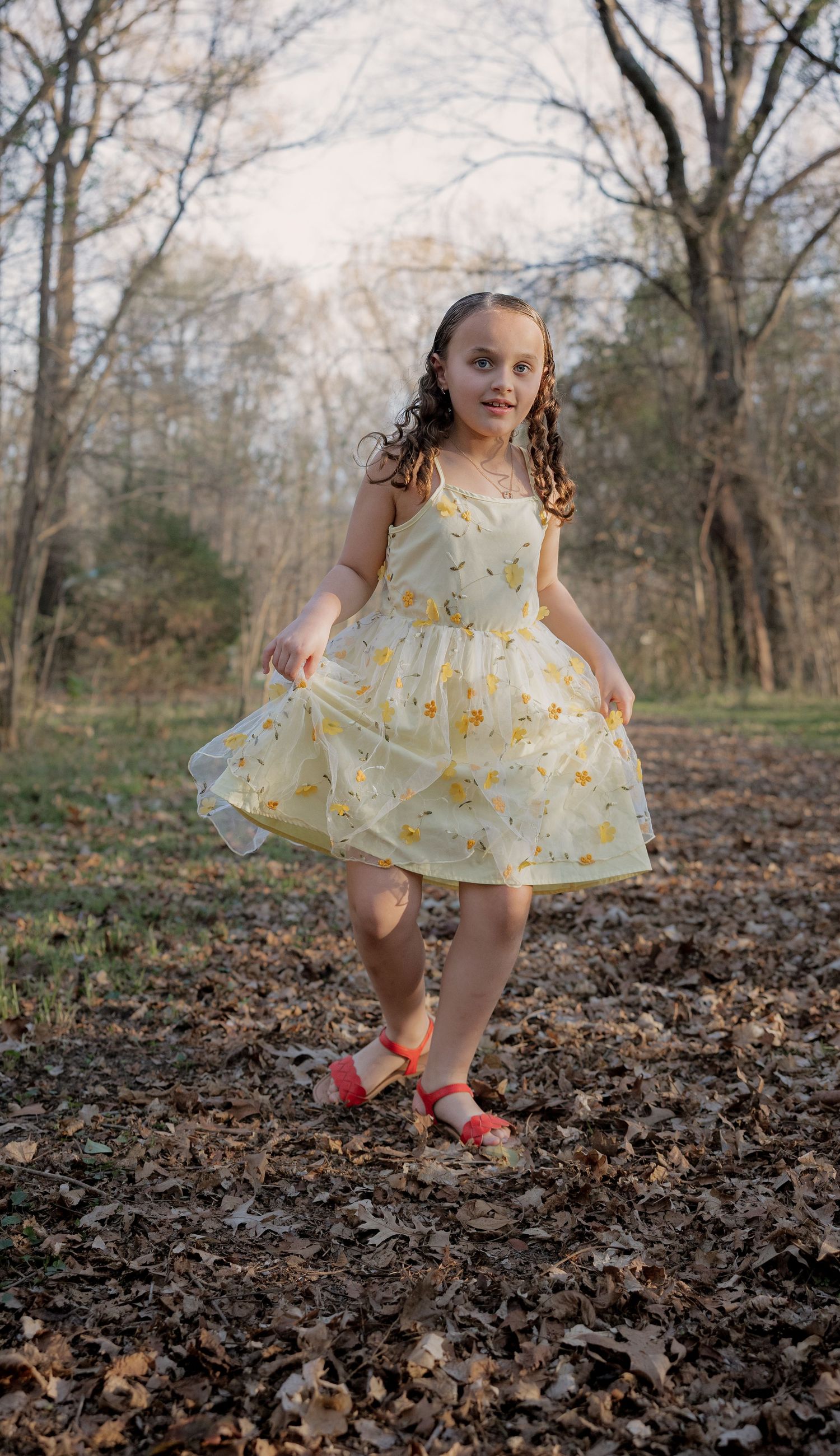 A young girl in a yellow floral dress and red shoes twirls among fallen leaves in a wooded autumn setting.