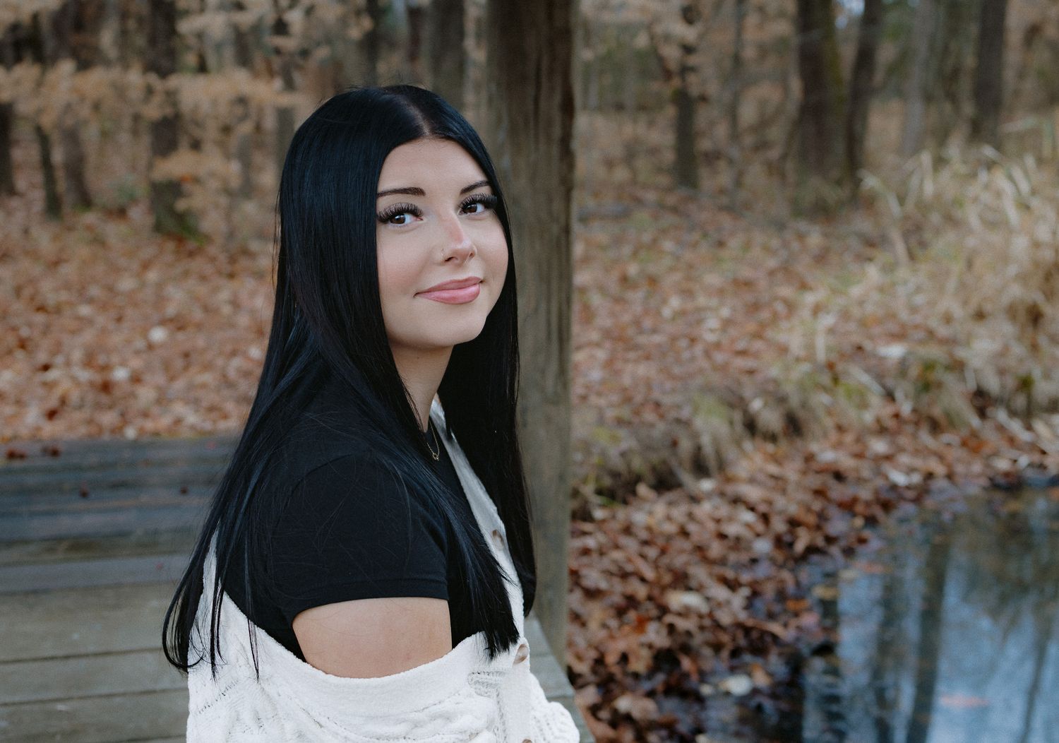 A person with long dark hair poses in a white sweater among autumn leaves during a fall photoshoot.