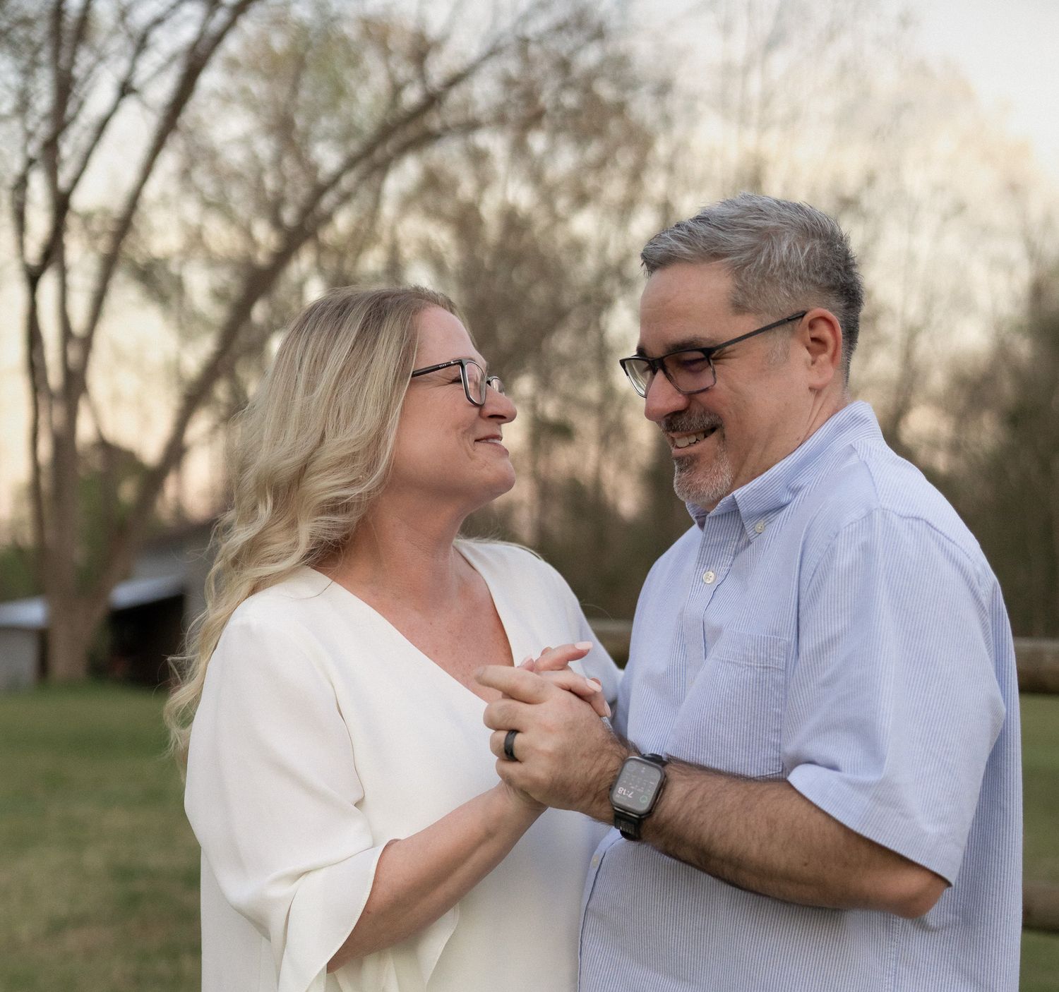 A middle-aged couple holds hands and smiles at each other outdoors, with trees and a barn in the background.