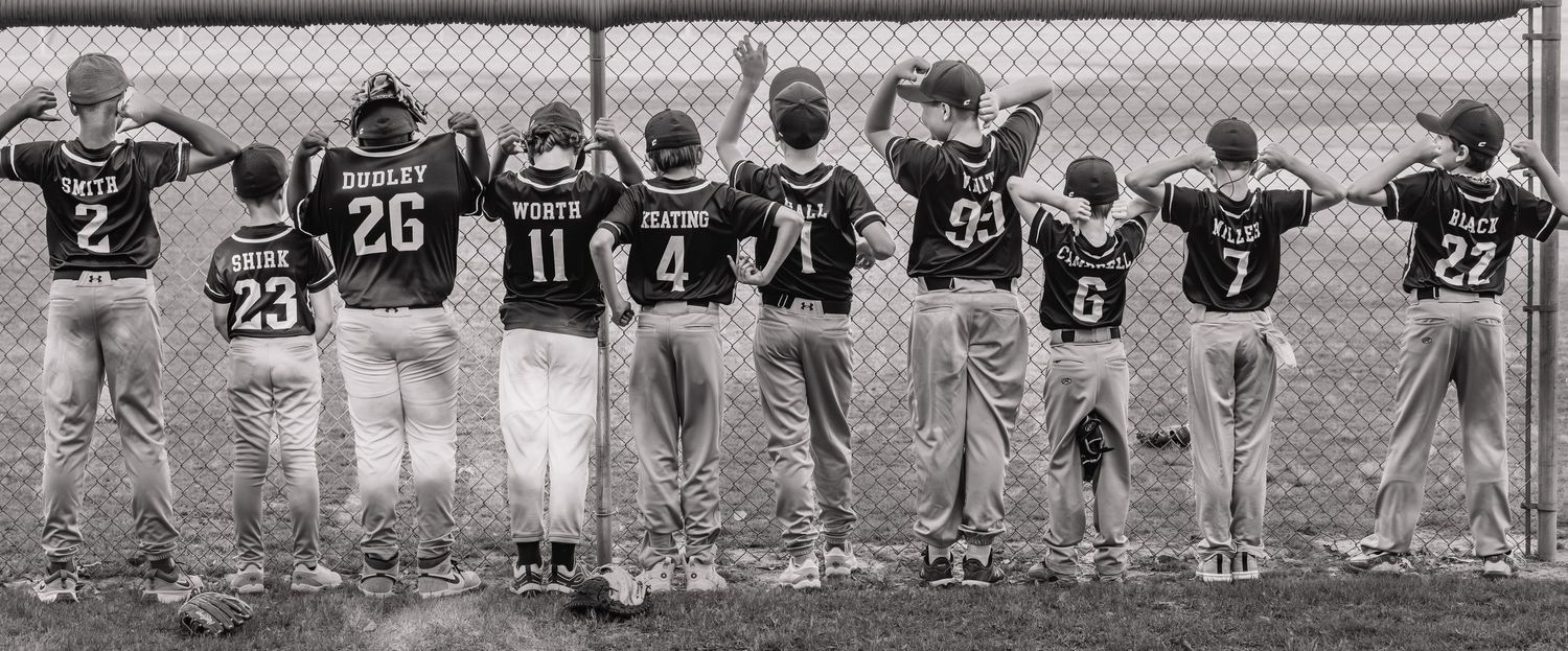Youth baseball team players standing together at a chain-link fence, viewed from behind, wearing numbered jerseys.