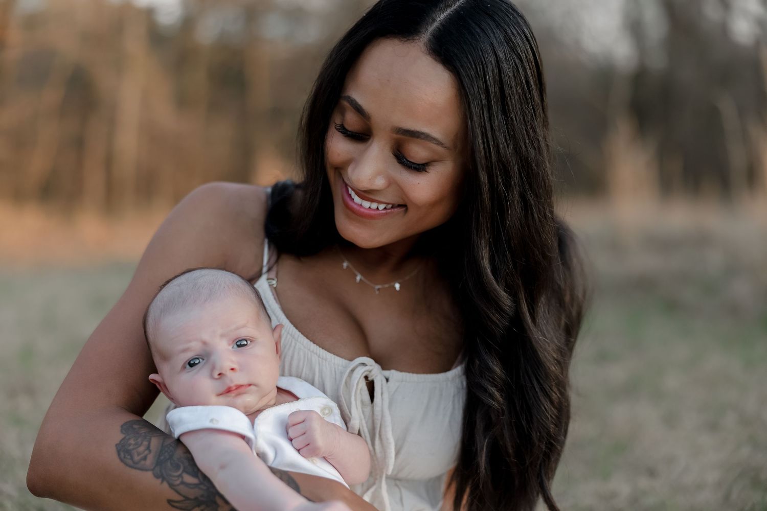A smiling young mother with long dark hair holds her newborn baby outdoors in a golden hour field setting.