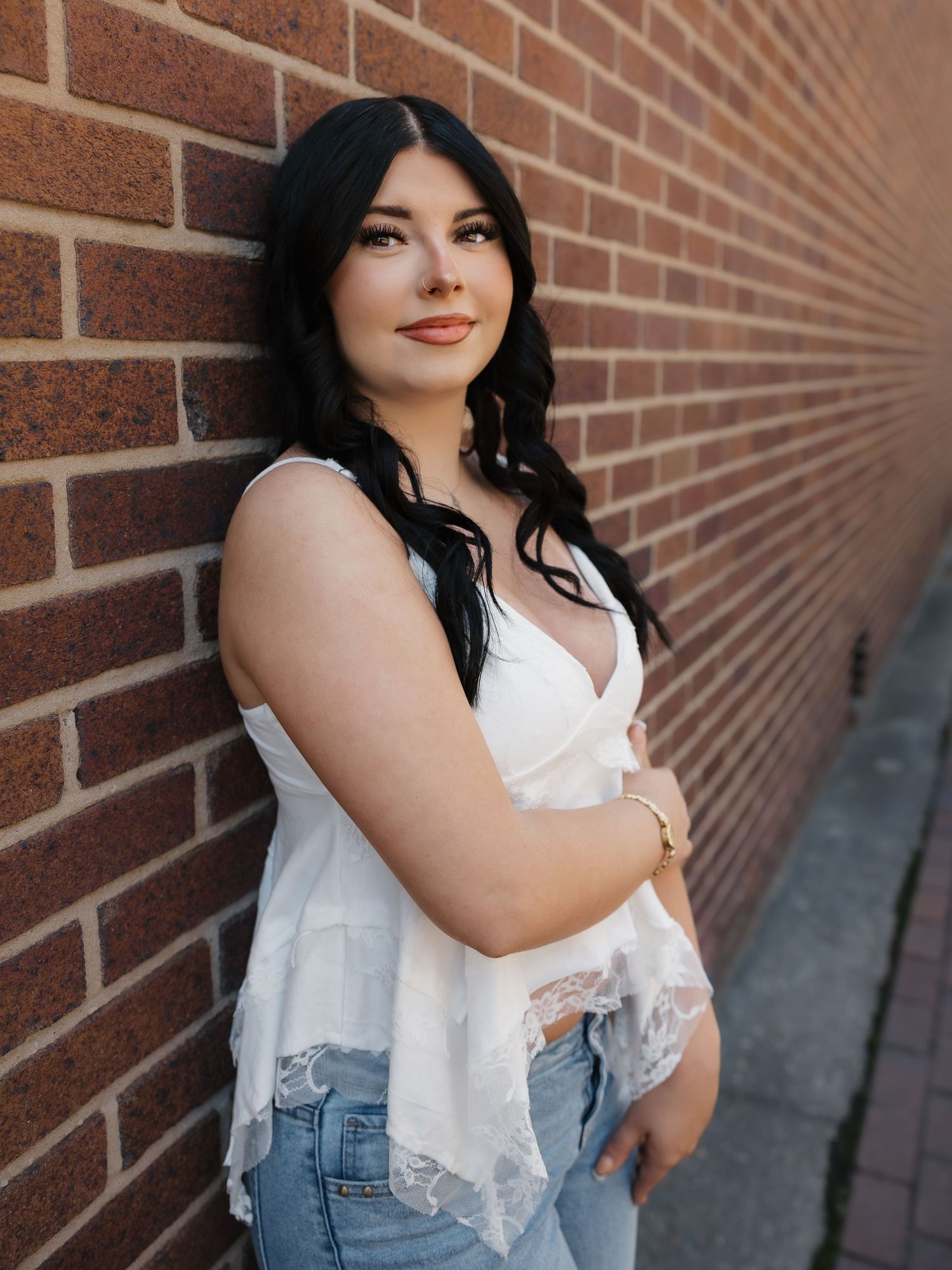 Young woman with dark hair in white lace top and jeans leaning against a brick wall outdoors.