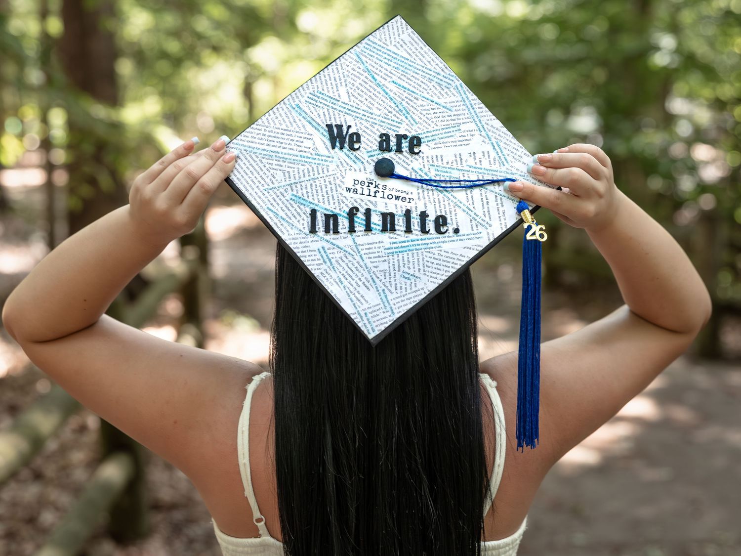 Graduate holds decorated cap reading 'We are infinite' with blue tassel in a forest setting.