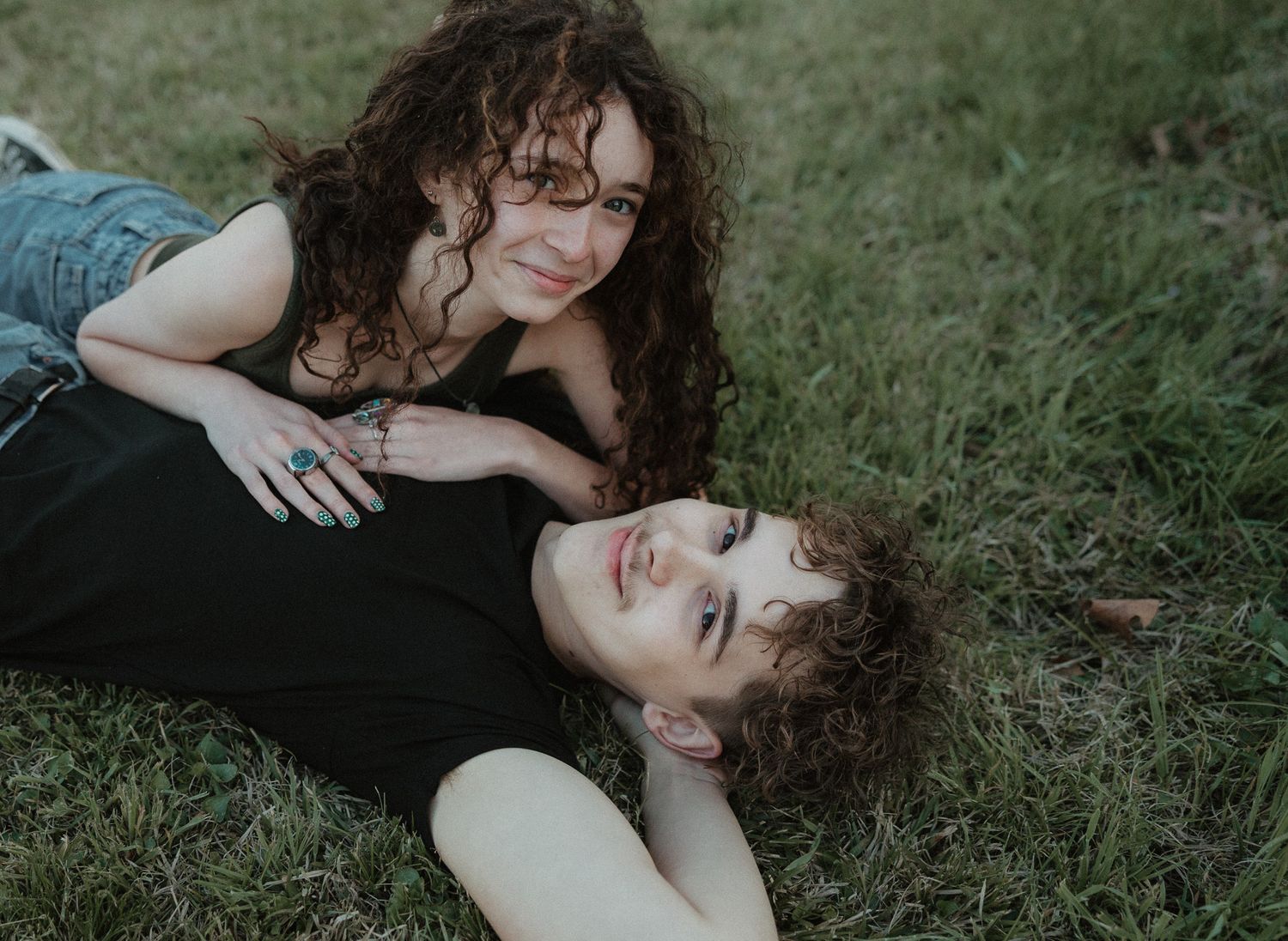 Two young women with curly hair relaxing on green grass, one lying down while the other leans over her smiling.