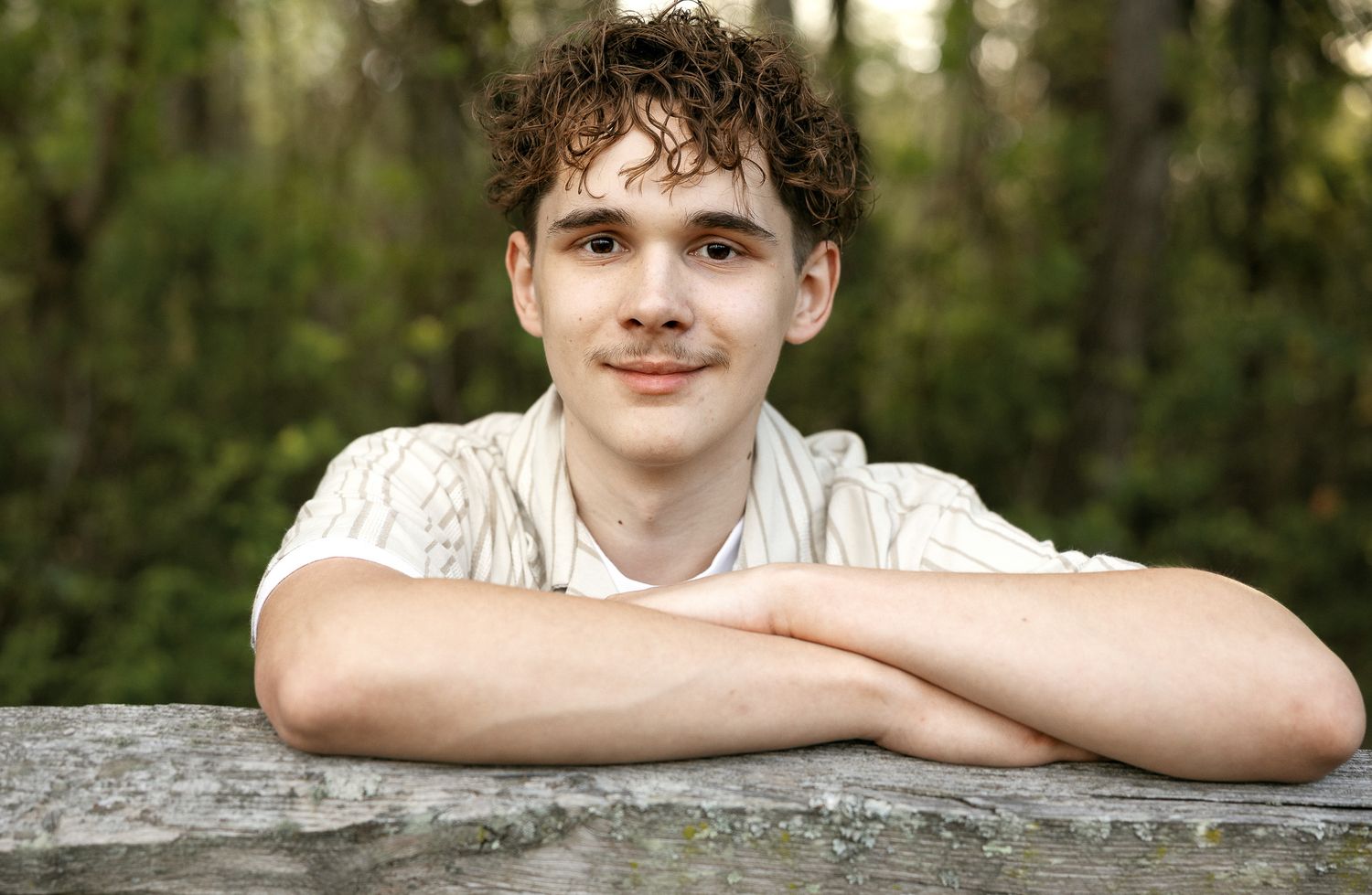 Young man with curly hair smiling while leaning on a stone surface outdoors with green trees in background.