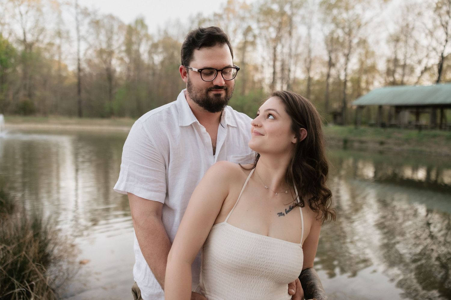 Couple posing romantically by a serene pond surrounded by trees during golden hour.