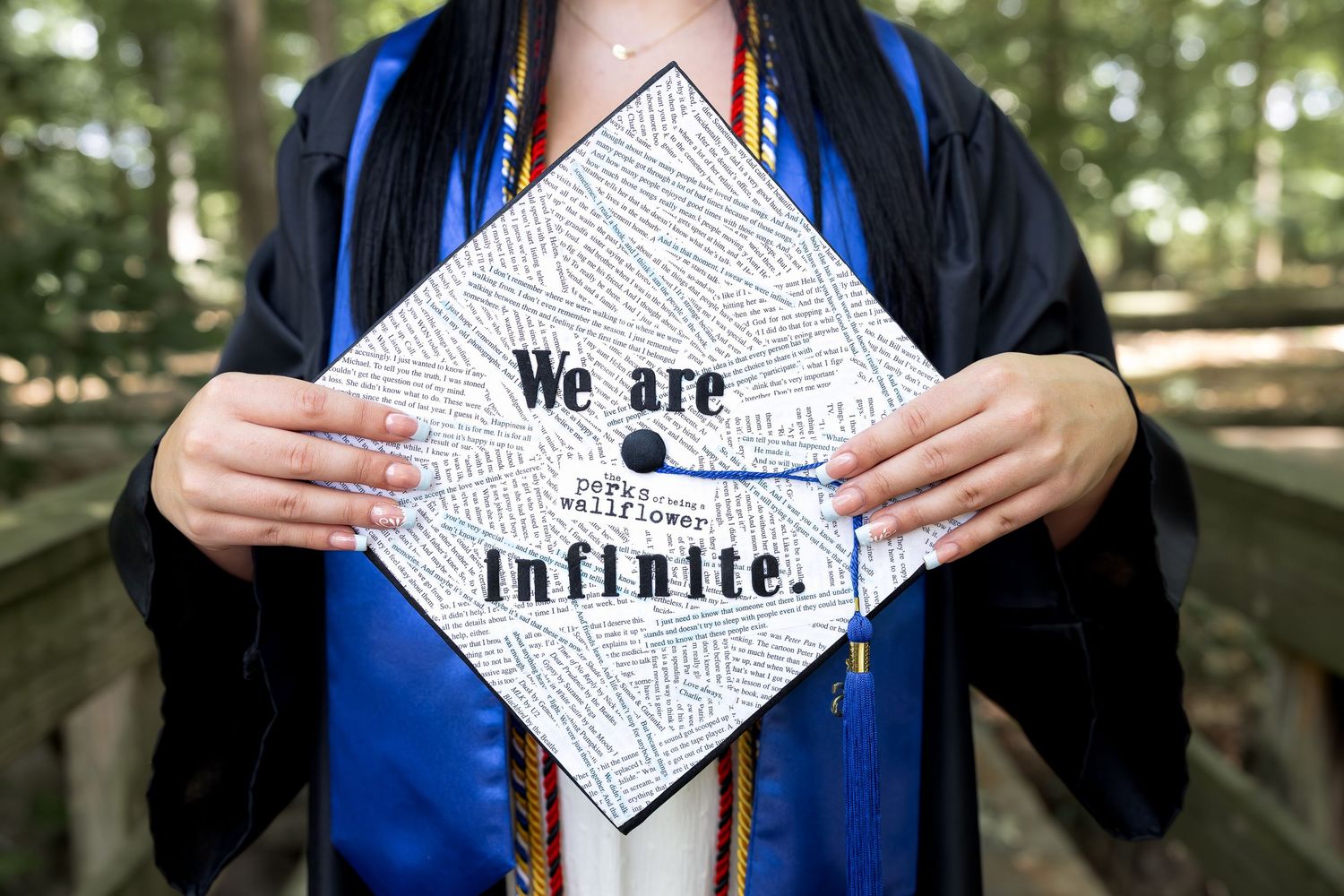 Graduate holding decorated cap reading We are infinite, a Perks of Being a Wallflower quote, wearing blue commencement gown.