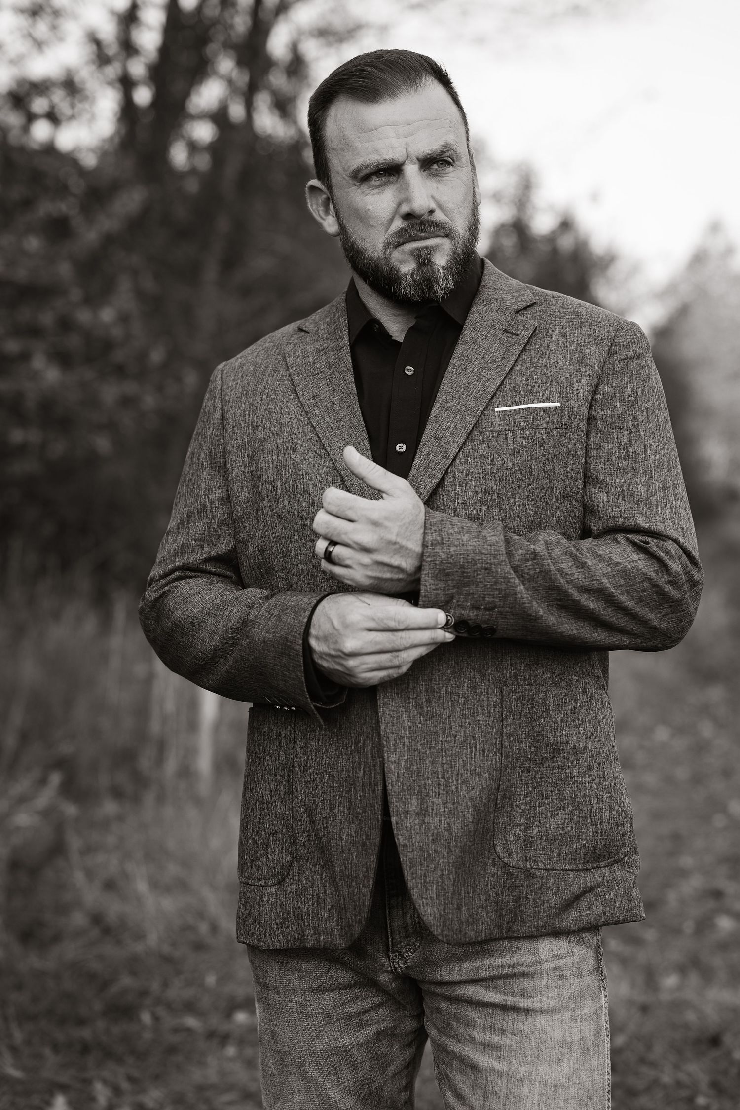 Man in tweed jacket and jeans poses thoughtfully in black and white outdoor portrait series.