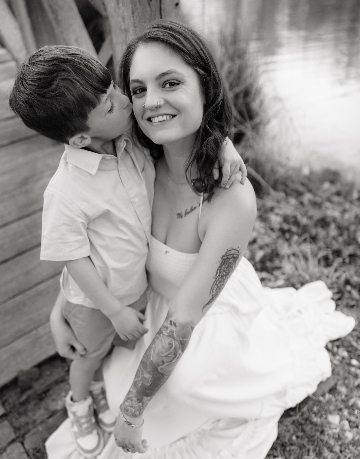 Black and white photo of a smiling woman with tattoos in a white dress hugging a young boy near a lake.