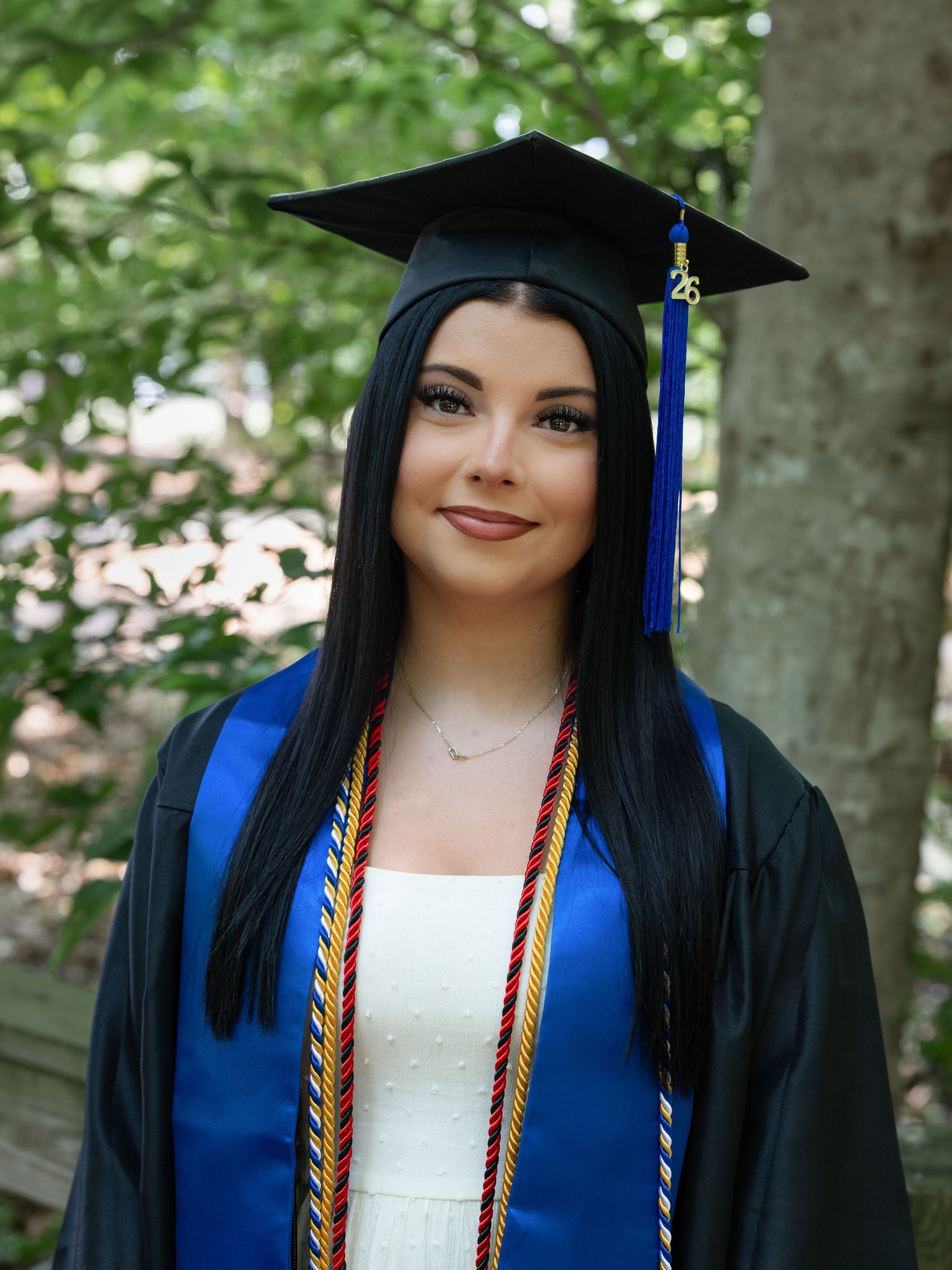 Young woman in graduation cap, gown, and blue stole smiles outdoors among green trees.