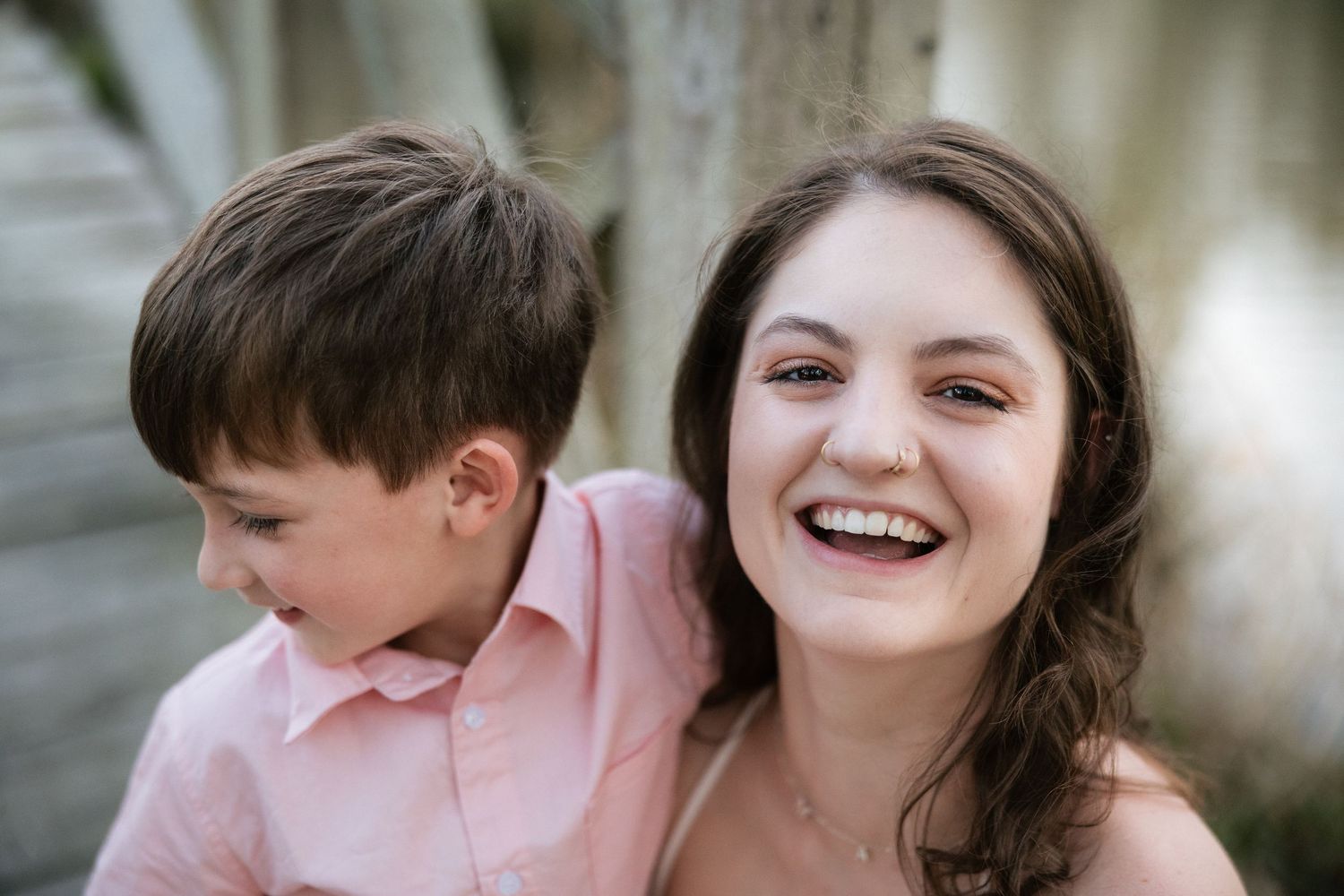 A joyful woman smiles brightly while a young boy in a pink shirt leans on her shoulder.