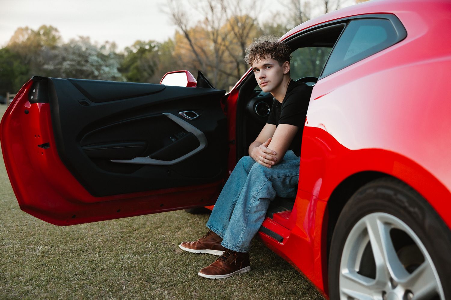 Young man in jeans and black shirt sitting in open door of red sports car on grass.