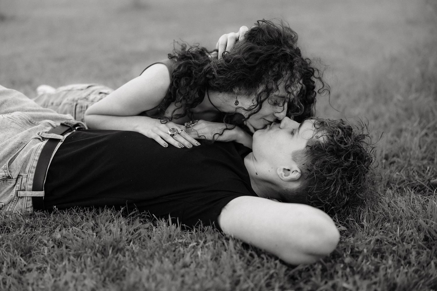 Black and white photo of a romantic couple lying on grass, sharing a tender kiss outdoors.