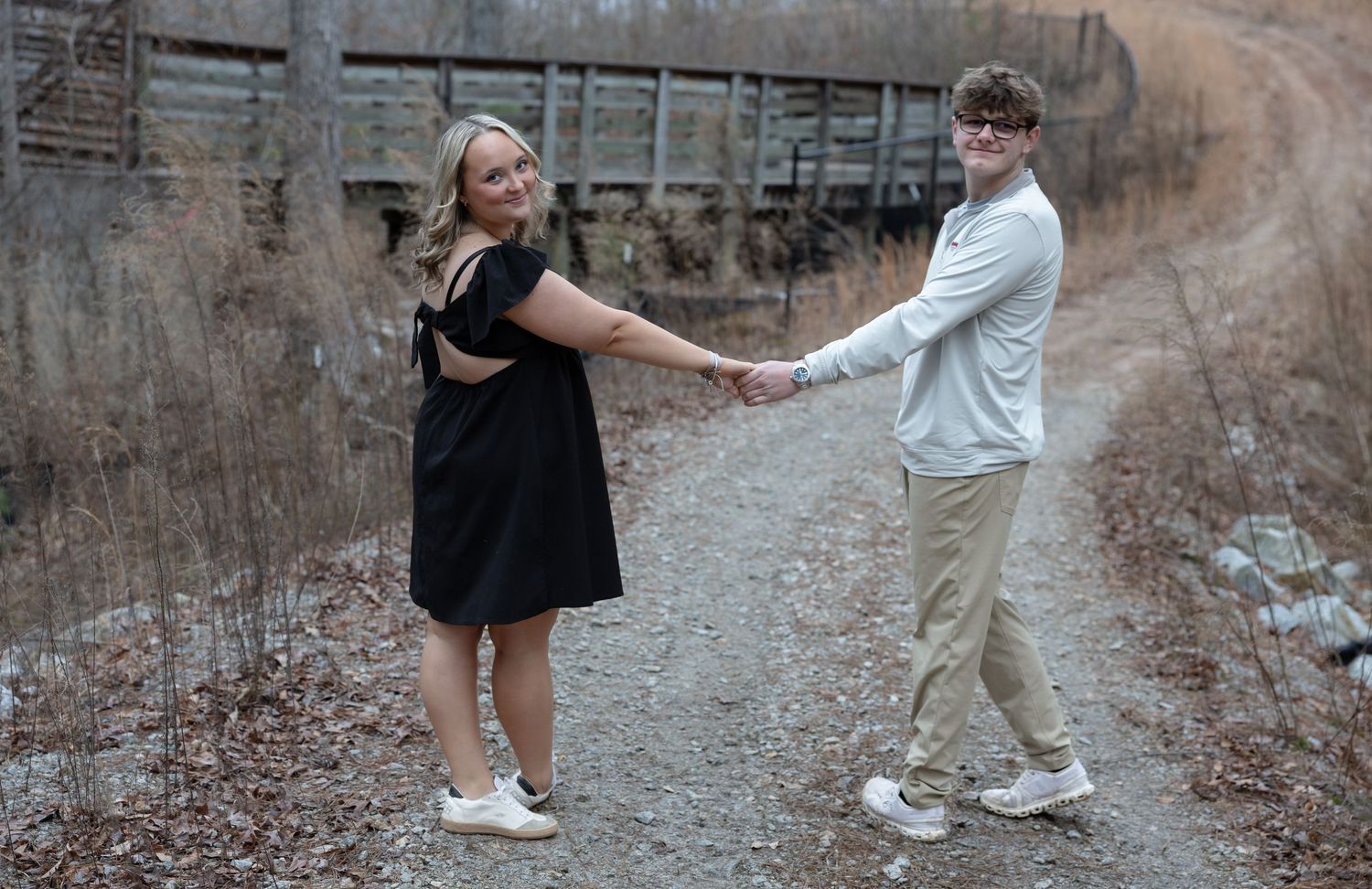 Couple holding hands on a gravel path outdoors, woman in black dress and man in tan outfit, with a bridge backdrop.