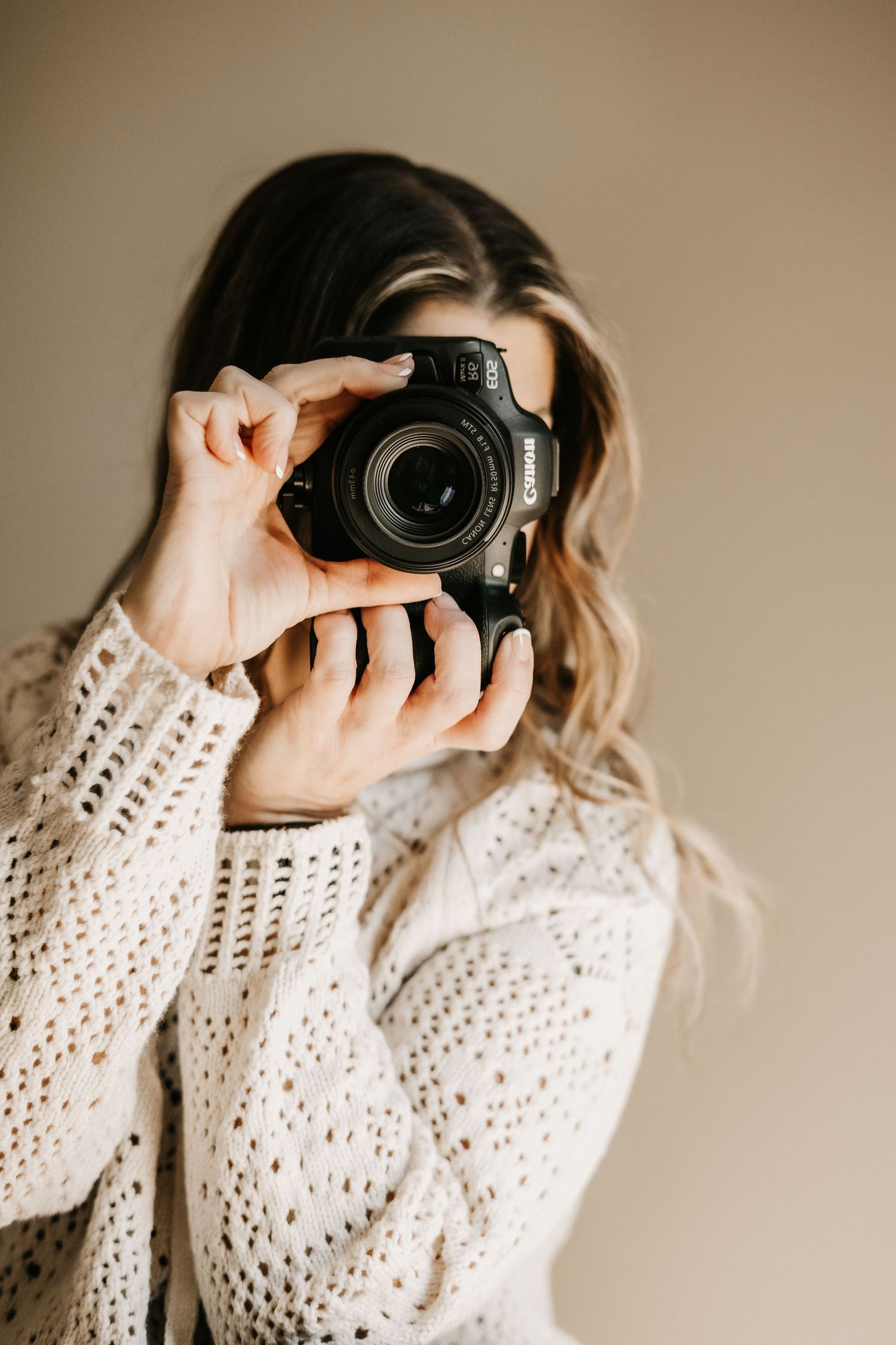 A photographer wearing a cream crochet sweater takes photos against a warm beige backdrop.
